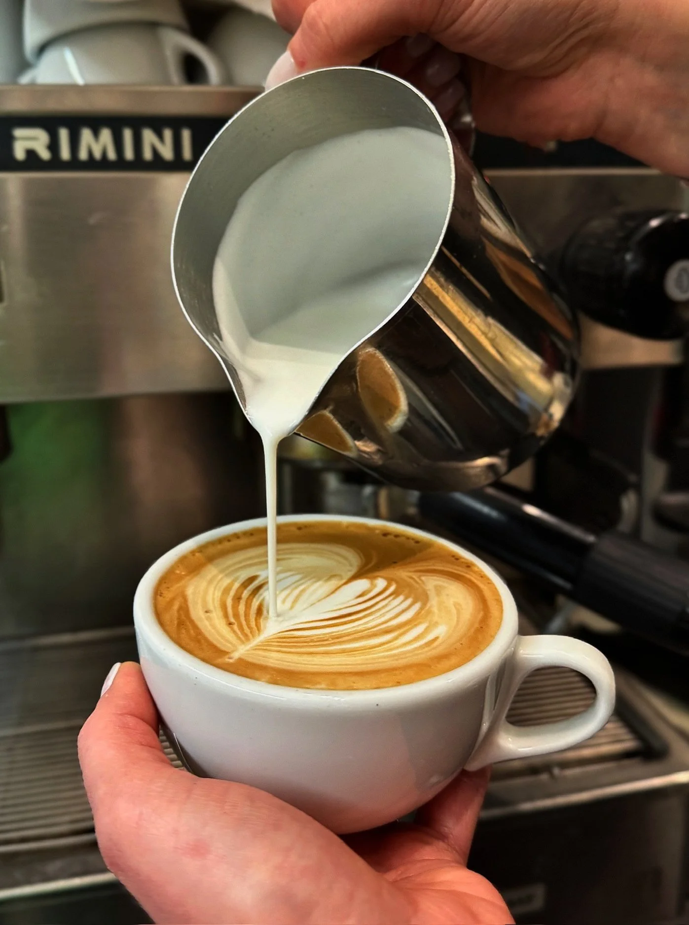 A barista pours steamed milk into a latte, creating latte art in a white ceramic cup.