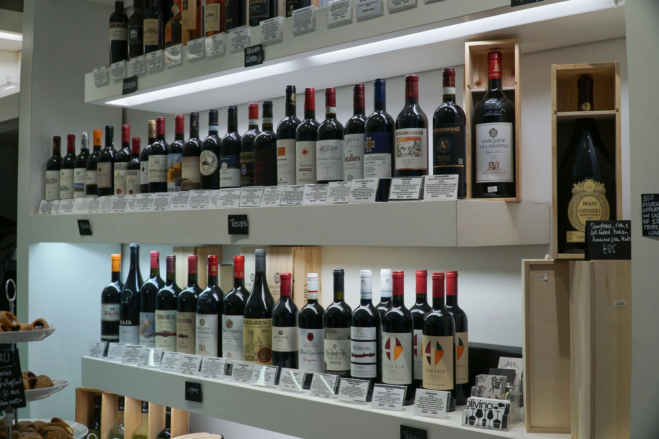 A well-organized display of various bottles of red wine on white shelves, with labels and small information tags underneath, situated in a retail store.