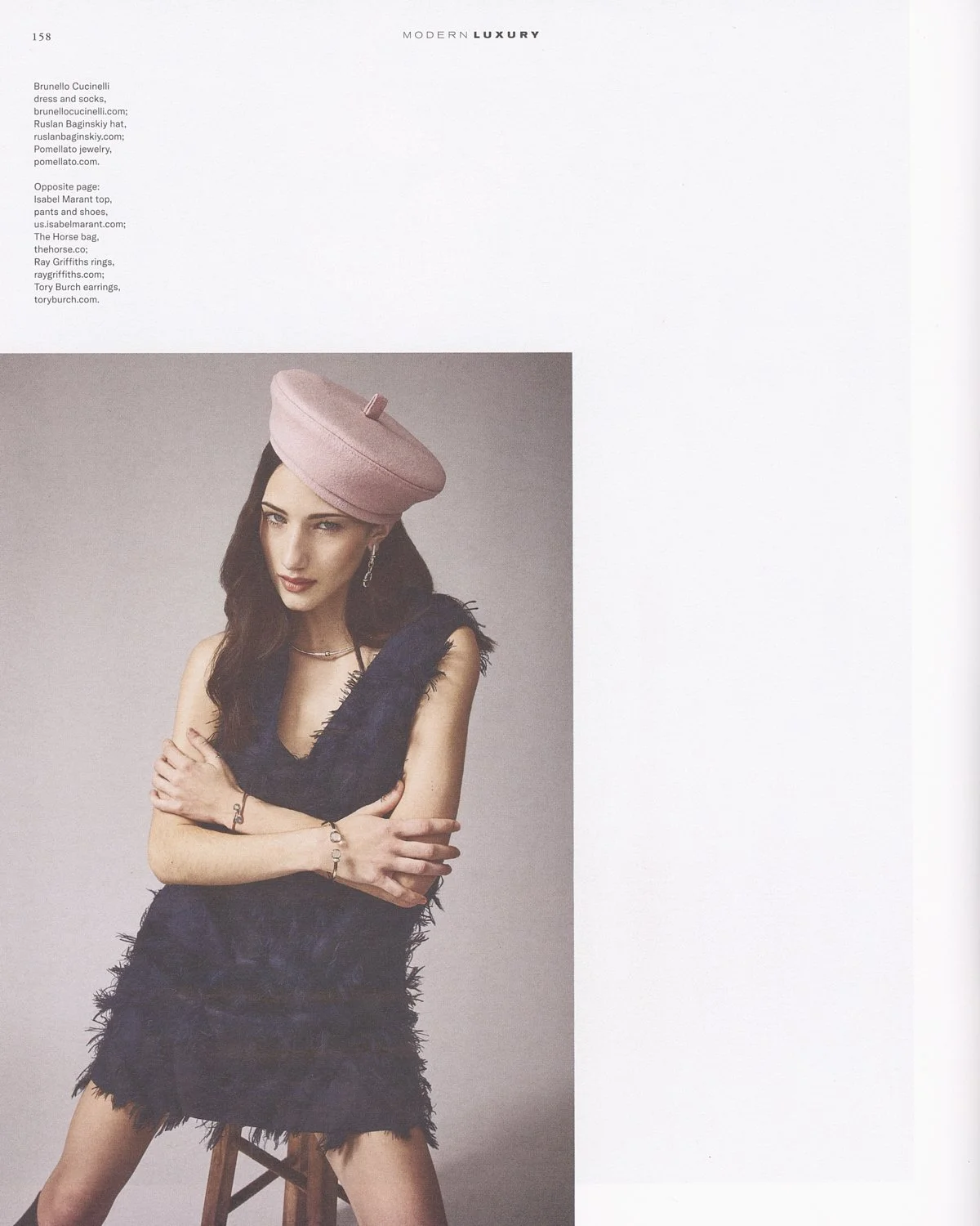A woman with dark wavy hair wearing a pink hat with a small bow, a dark fringed dress, earrings, a necklace, and bracelets, sitting on a wooden stool with her arms crossed over her chest against a plain background.