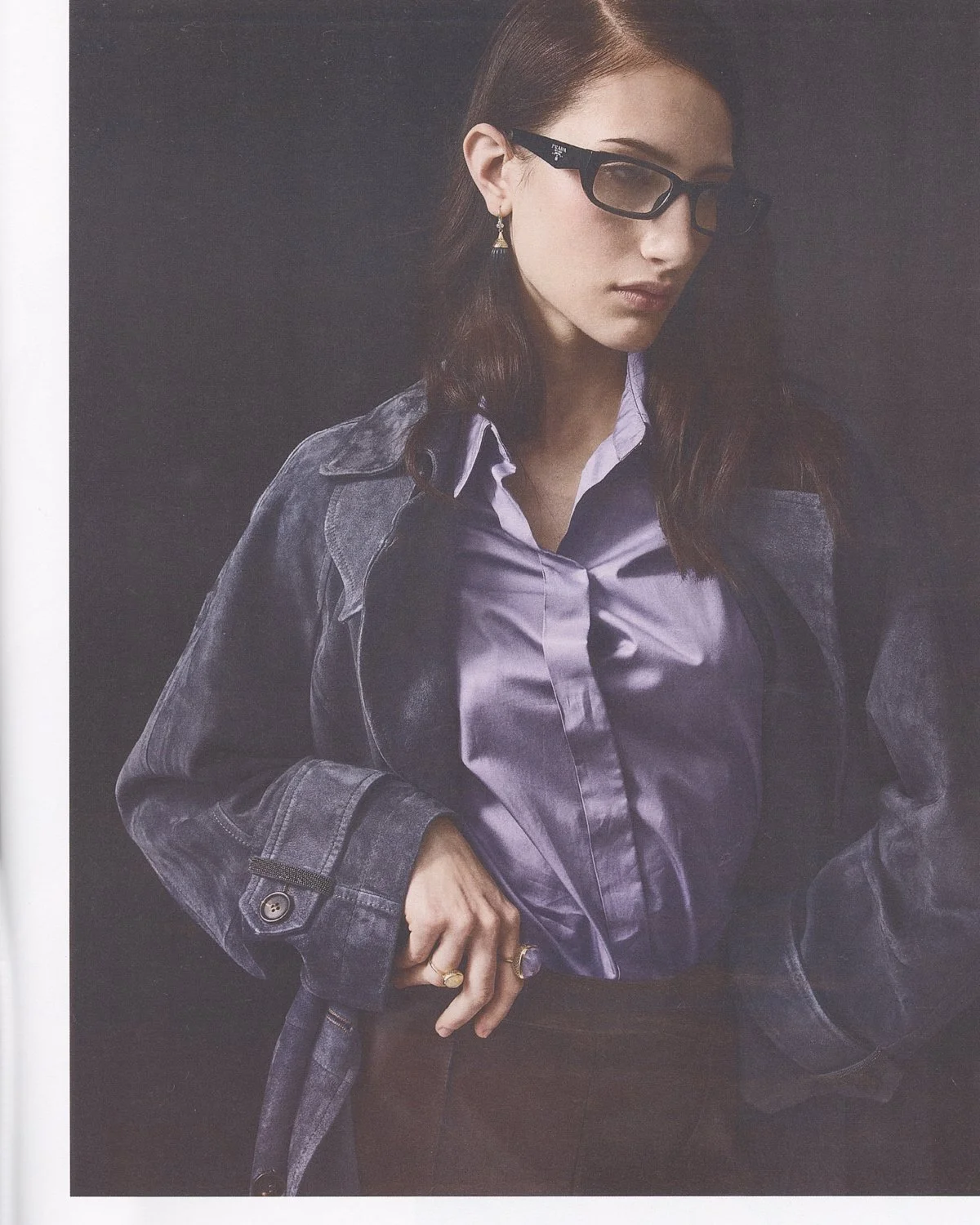 A woman with brown hair and glasses, wearing a purple satin shirt and a gray denim jacket, with her hand on her waist.