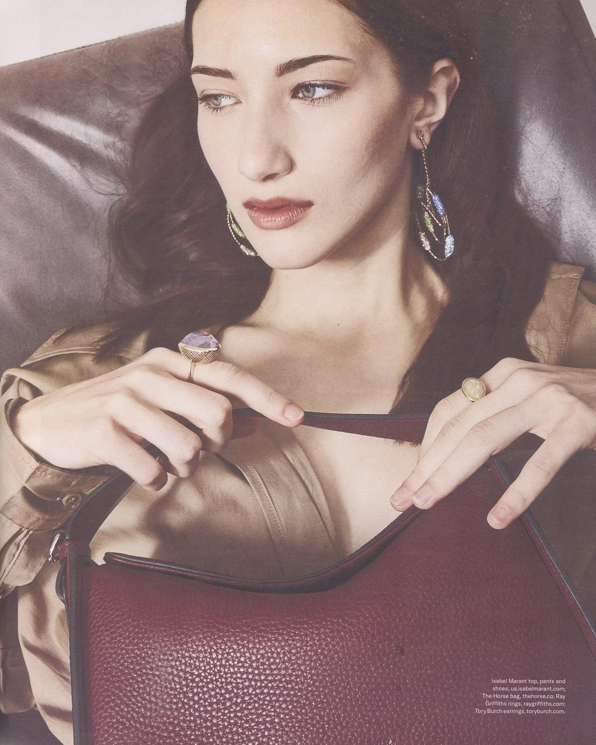 A woman with dark, wavy hair, red lipstick, and earrings, holding a large red handbag with her right hand and a ring on her left hand, seated on a leather couch.