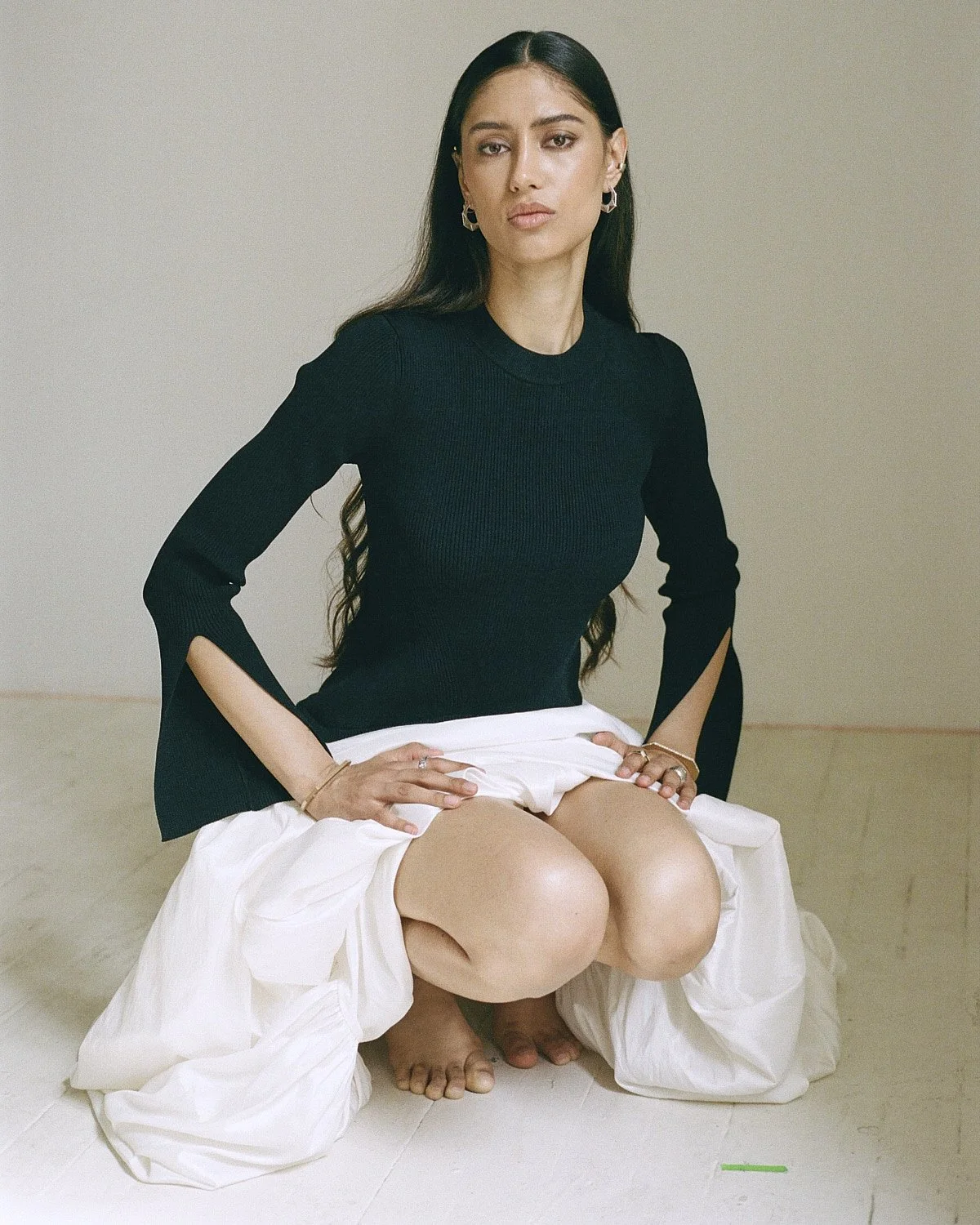 A woman with long dark hair, wearing a black top and a white skirt, kneeling on the floor with her hands on her thighs, looking at the camera against a plain background.