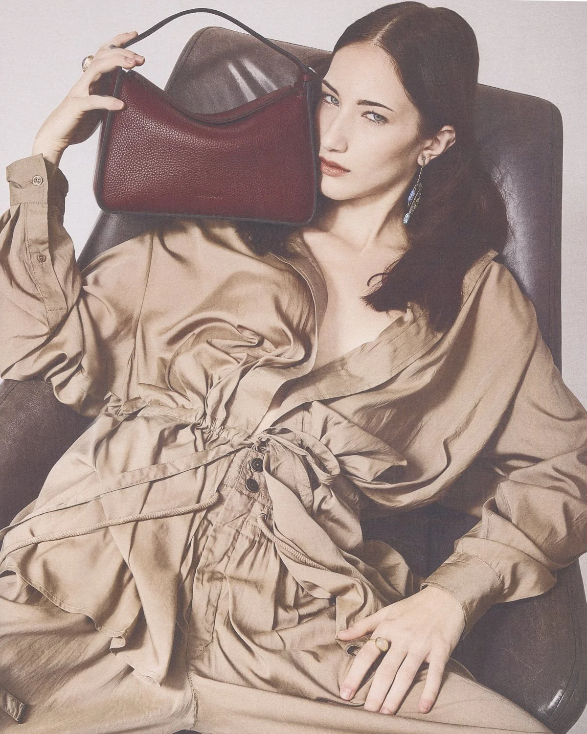 A woman with dark hair, wearing a beige dress, sitting on a brown chair, holding a red handbag on her shoulder, with one eye half-closed.