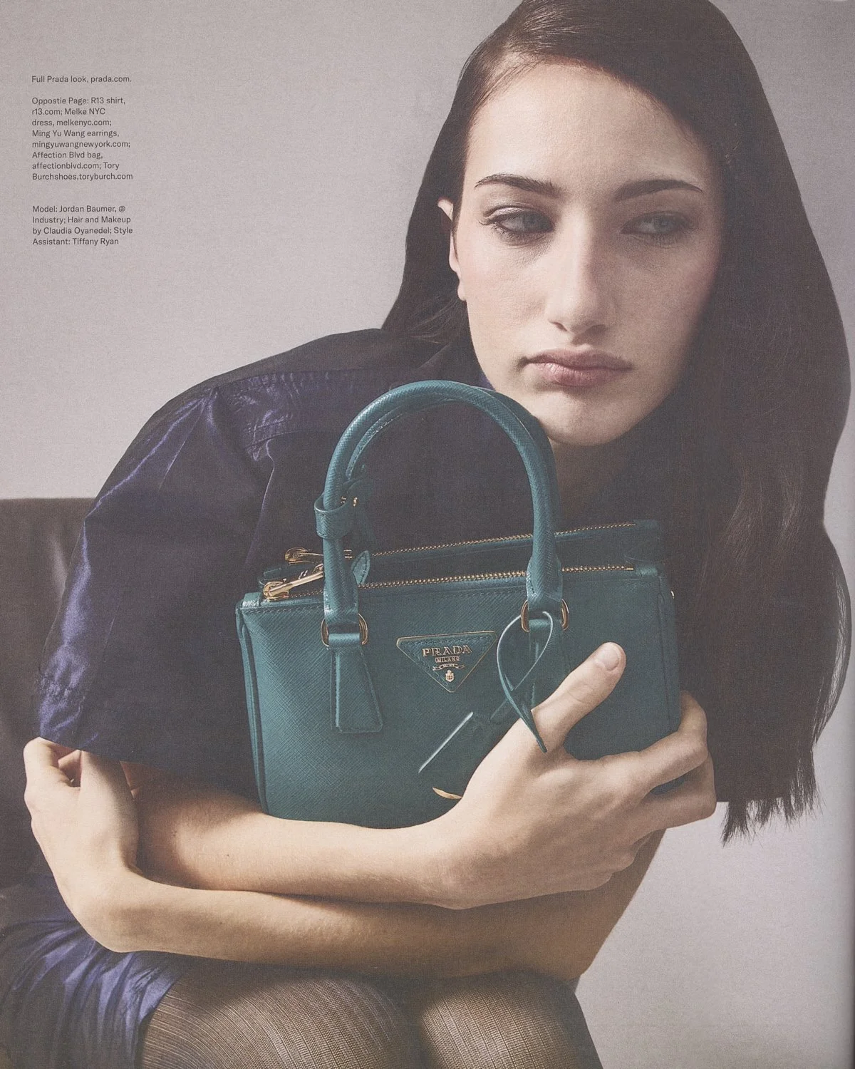 A woman with dark hair and fair skin holds a teal Prada handbag while sitting on a chair. She is dressed in a black leather jacket and looking slightly to the side with a neutral expression.