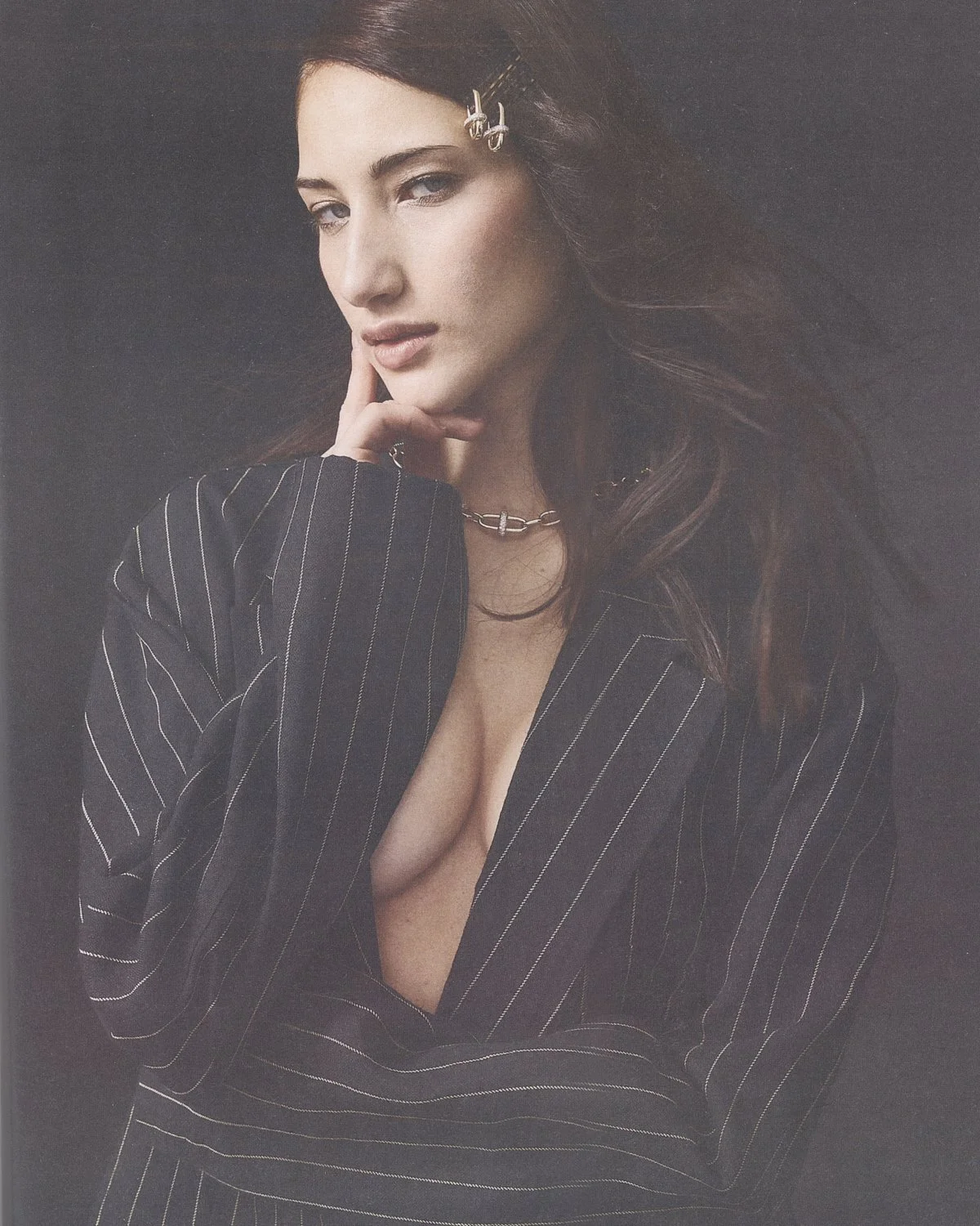 A woman with long, dark hair and fair skin, dressed in a black and white pinstripe blazer, poses with her hand touching her face against a dark background.