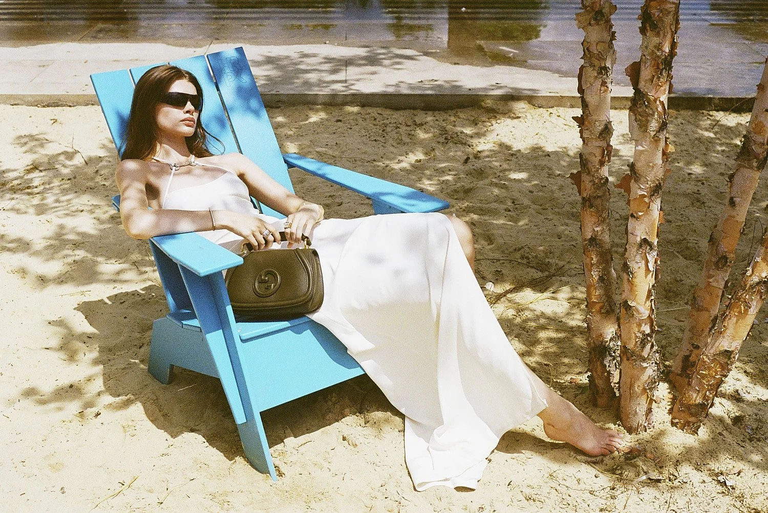 Lexie Tapper in sunglasses and a white dress is relaxing on a blue Adirondack chair on a sandy beach, holding a black handbag on her lap. There are tree trunks nearby, and sunlight filters through the leaves.
