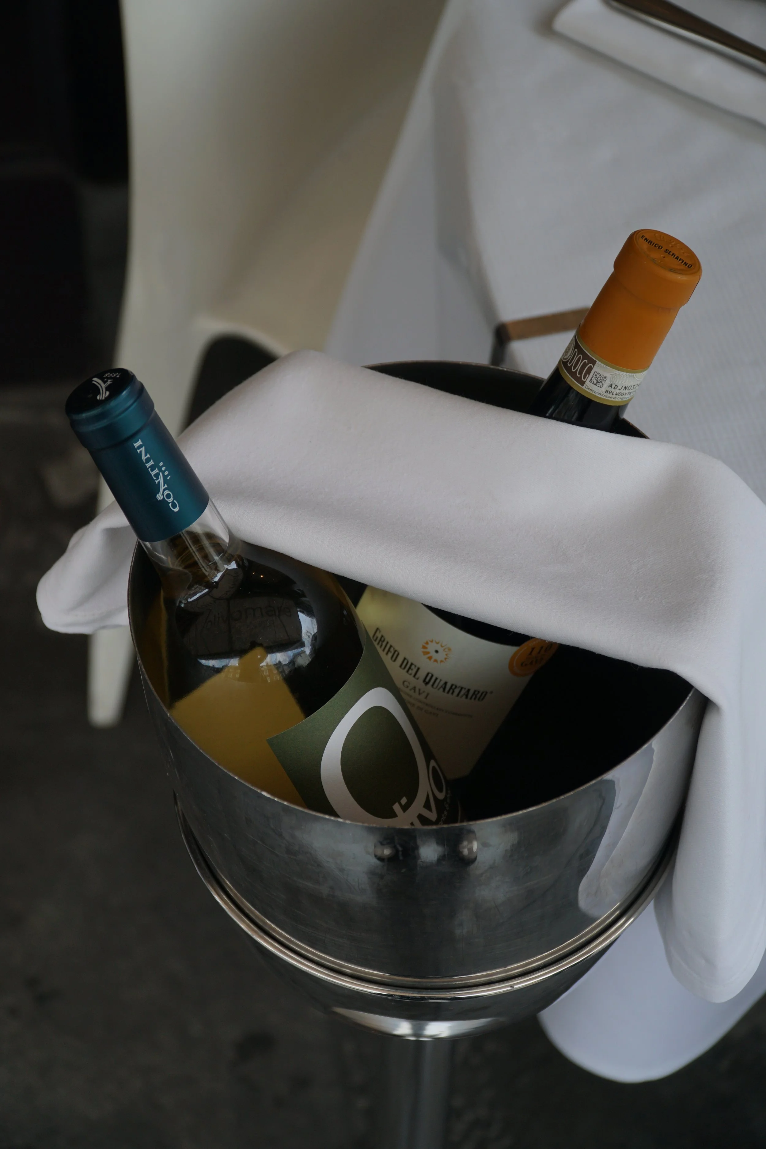A metal ice bucket holding two bottles of wine with one covered by a white cloth, on a table near white chairs in a restaurant or event setting.