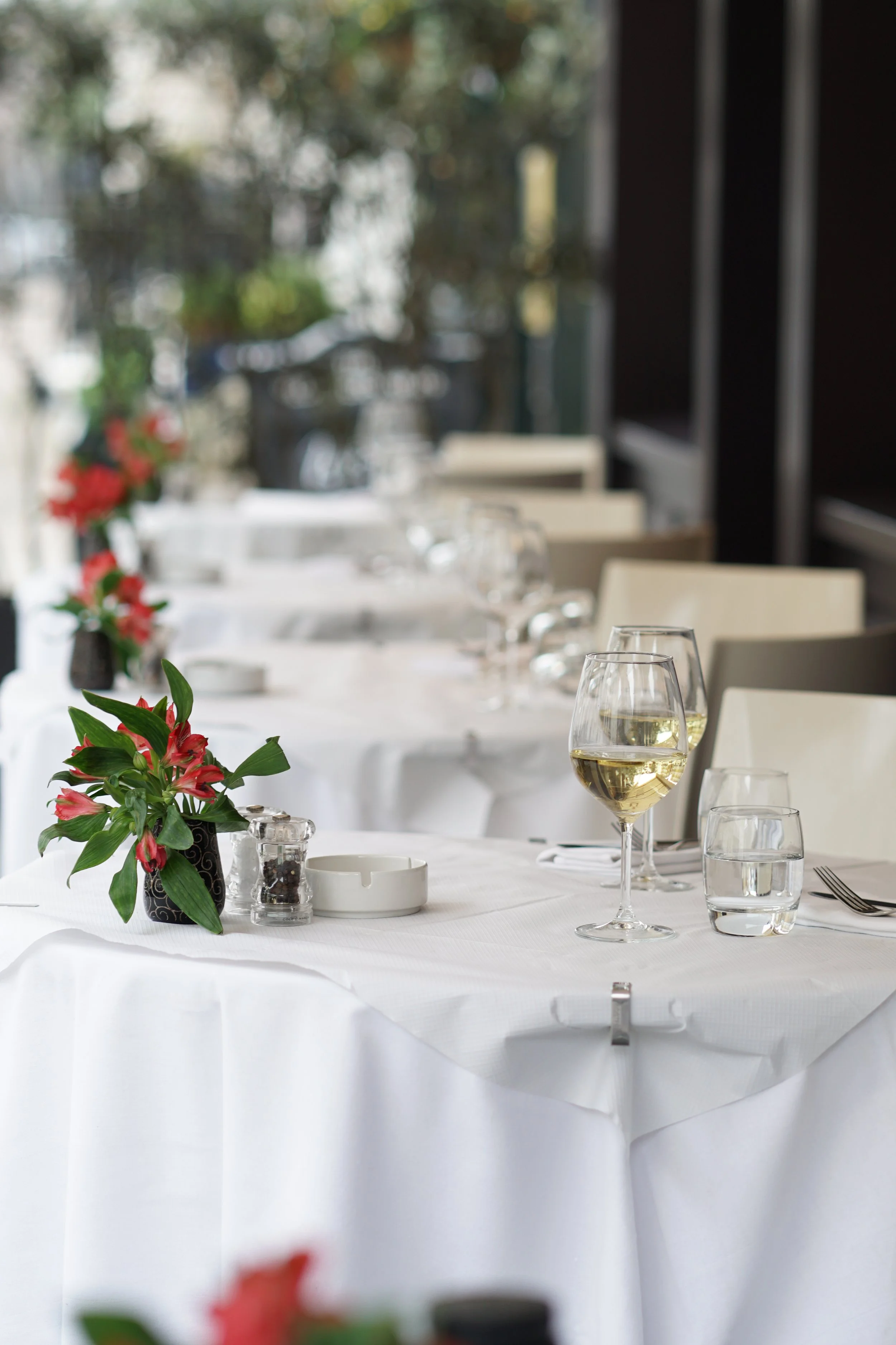 A long dining table set with white tablecloths, wine glasses with white wine, water glasses, floral centerpieces with red flowers and green leaves, and cutlery in a bright, elegant restaurant or dining area.