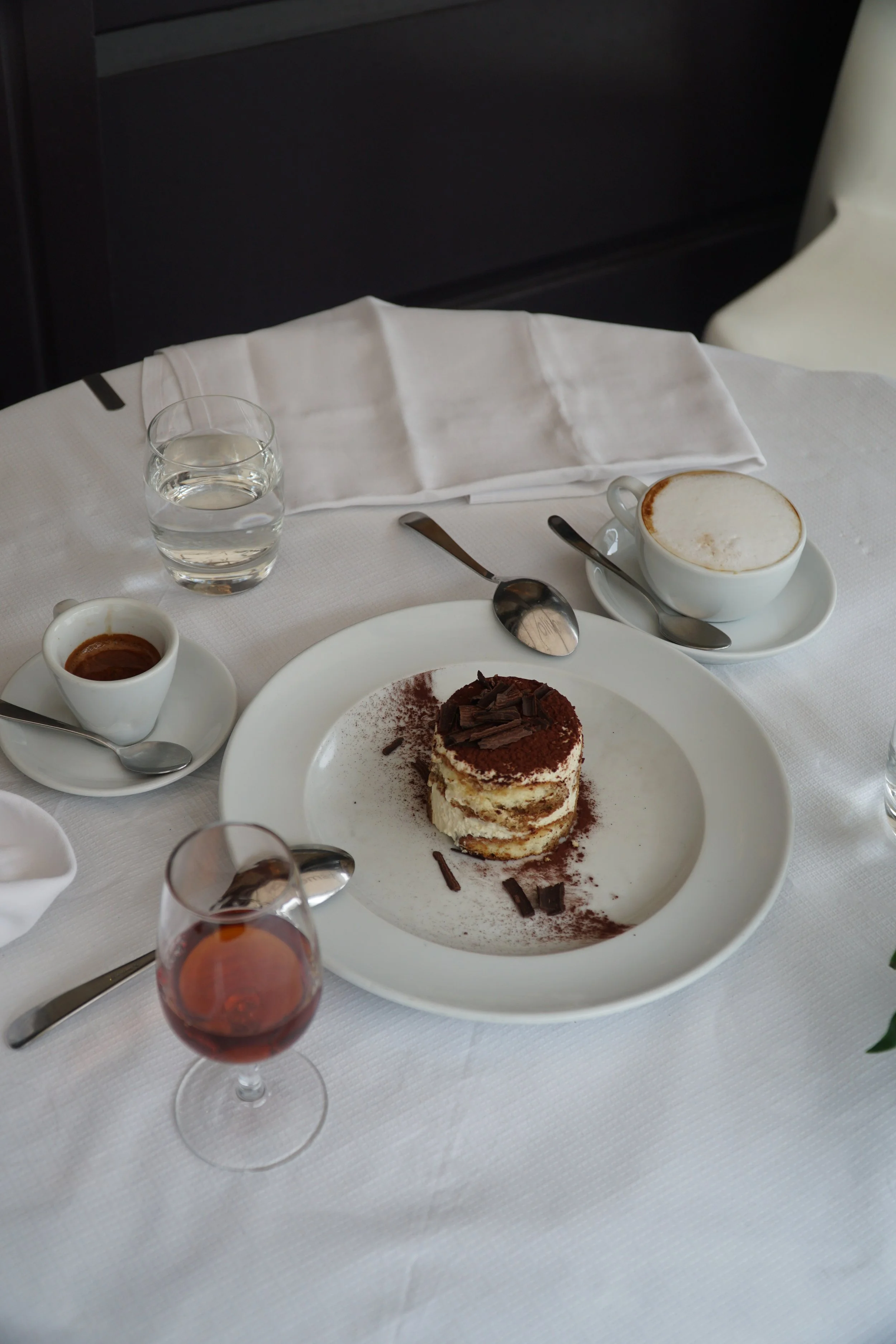 A white plate with a small layered tiramisu dessert topped with chocolate shavings and cocoa powder, surrounded by a cup of espresso, a cup of cappuccino, a glass of water, and a glass of red wine, all on a white tablecloth with silverware and white 