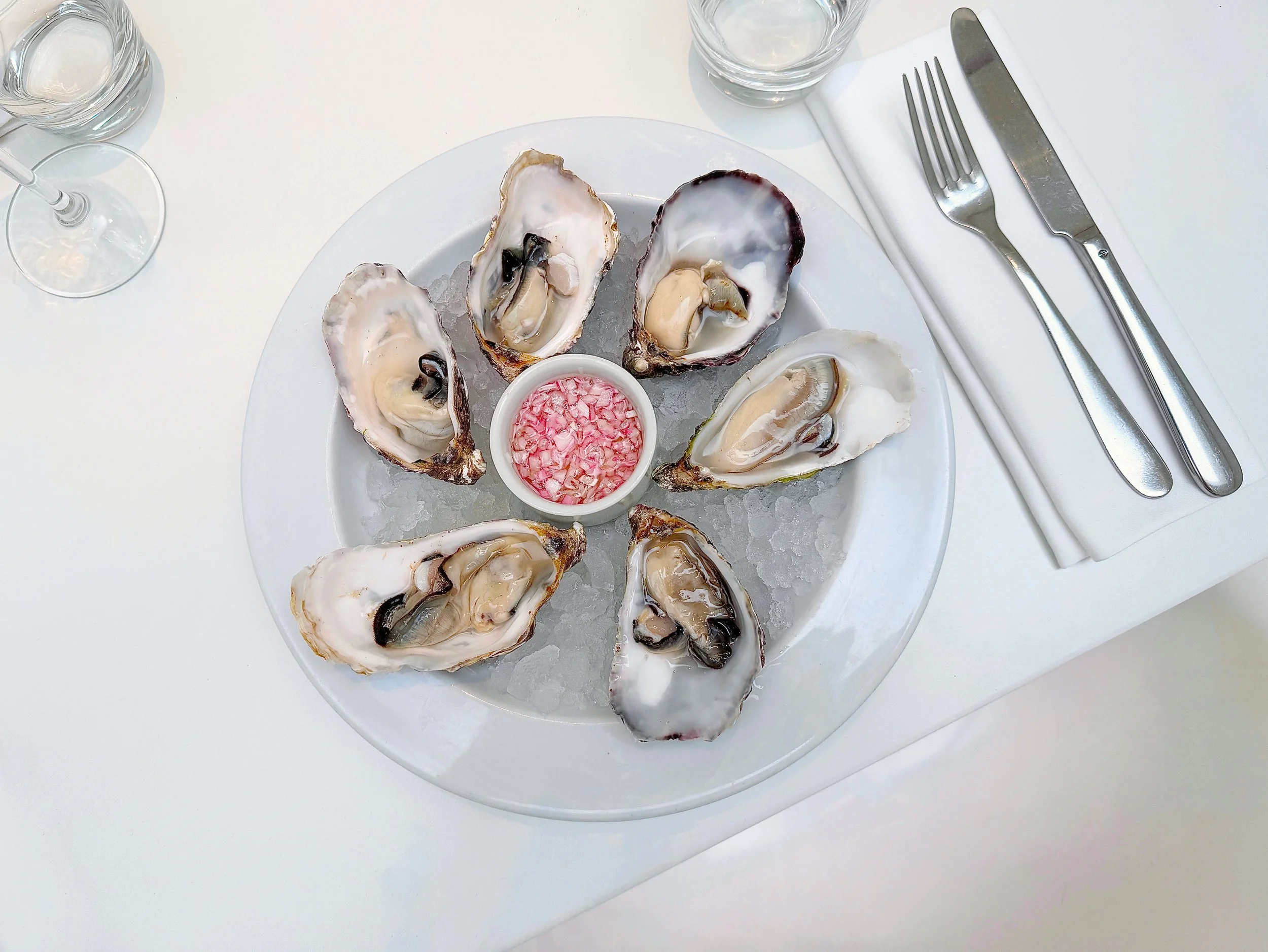 A white plate with several oysters on the half shell arranged around a small bowl of pink cocktail sauce, served on a bed of ice. There are two glasses, a fork, a knife, and a white napkin on the table.