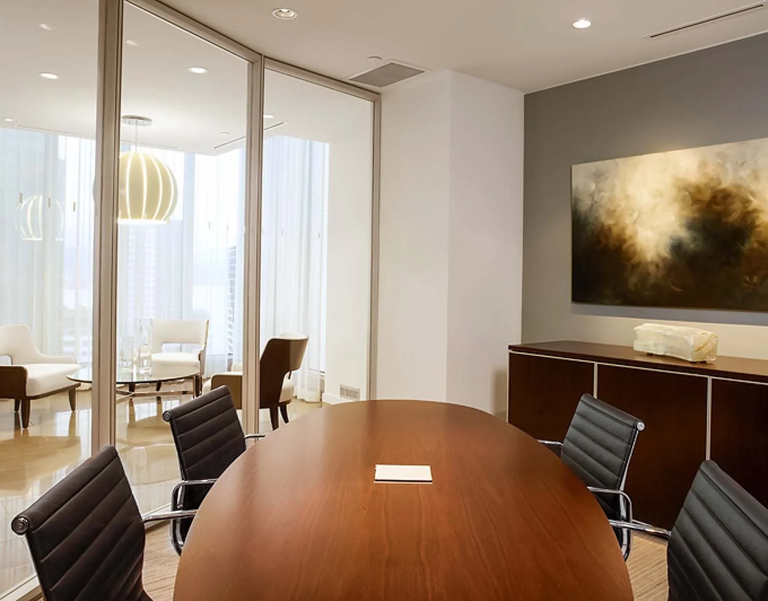 A modern conference room with a large wooden table surrounded by black leather chairs, facing a cabinet with a large abstract painting on the wall, and a glass partition leading to a lounge area with white armchairs, a small table, tall windows, and 