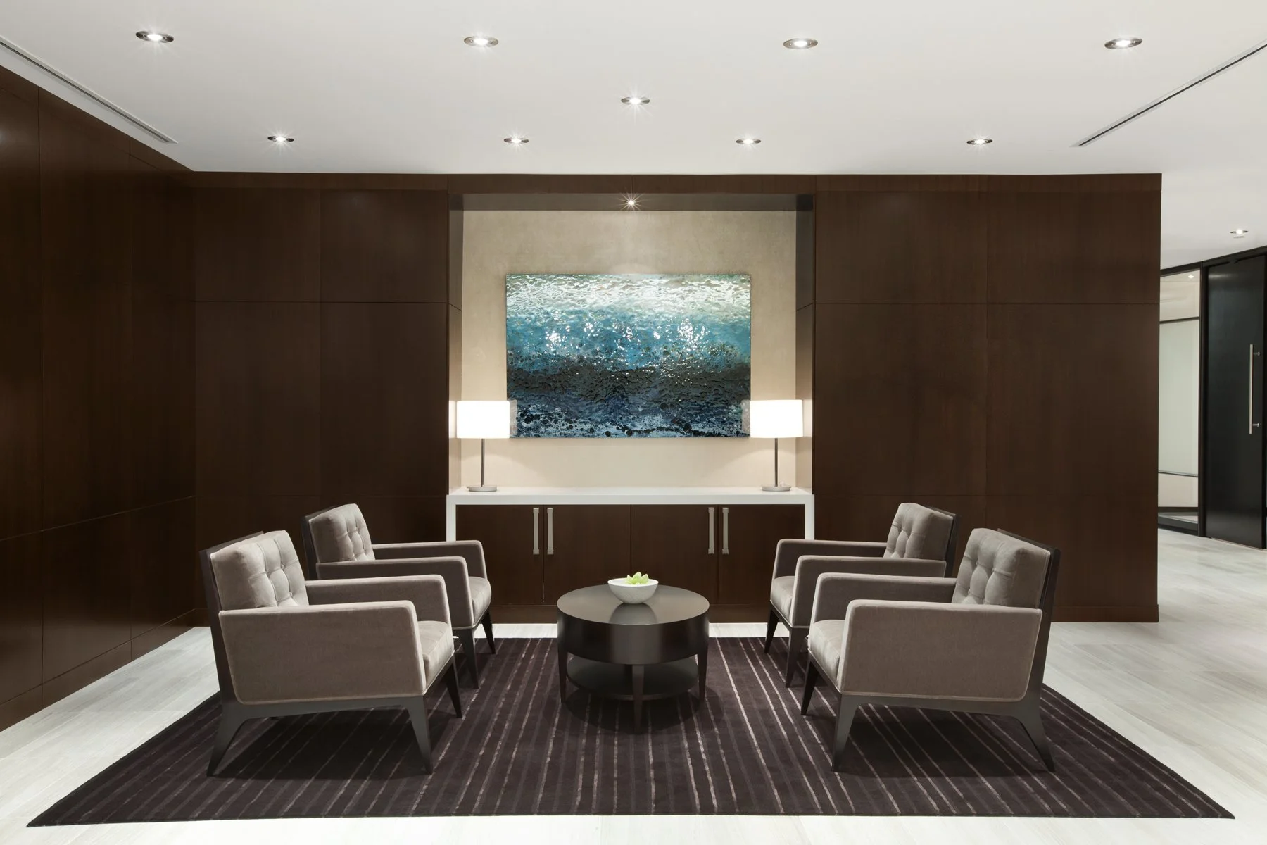 Modern waiting area with six gray upholstered chairs surrounding a black round coffee table on a striped rug. A white console with two table lamps and a large ocean-themed painting hangs on a beige wall in the background.