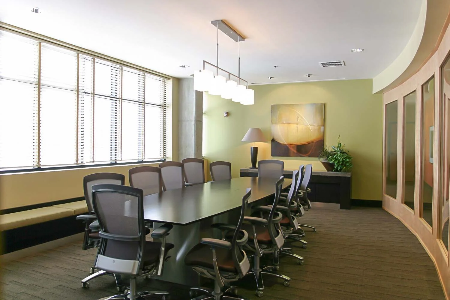 Modern conference room with a large table surrounded by office chairs, a large window with blinds, a wall with a painting and a plant, a sideboard with a lamp, and a curved wooden partition.