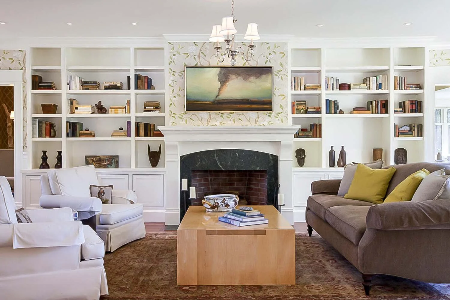 Historic living room with white built-in bookshelves, a fireplace with a black marble mantle, a wooden coffee table, and sofas with yellow and beige cushions. A framed painting of a smoky landscape hangs above the fireplace.