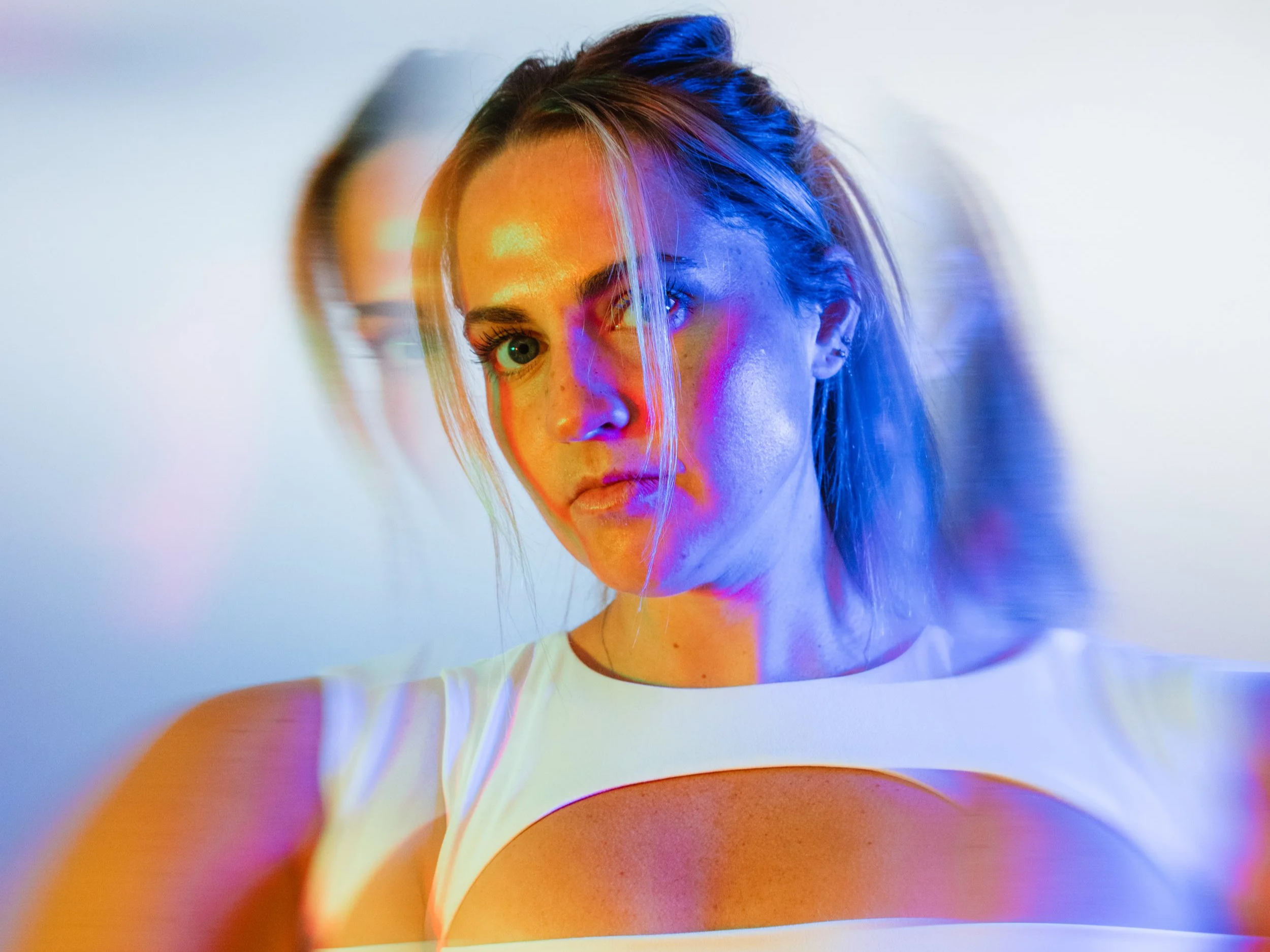 A woman with blonde hair and blue eyes looking at the camera, with colorful light effects creating a surreal visual with a split, mirrored effect.