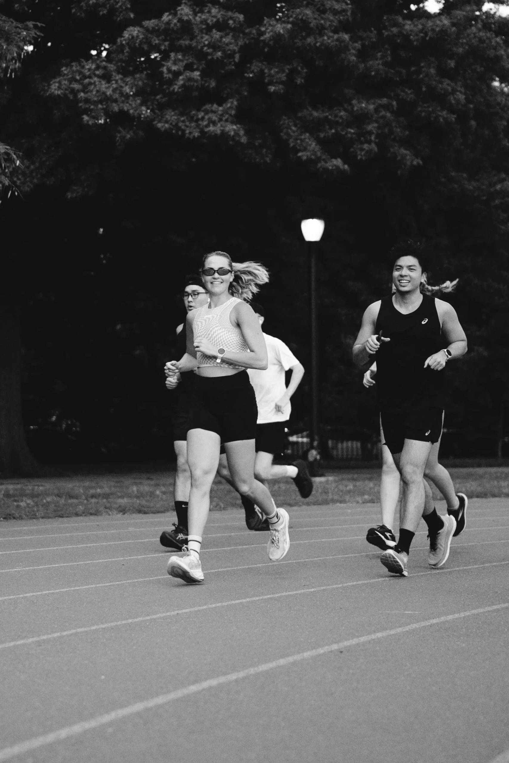 Group of runners running on an outdoor track at dusk, with trees and a lamp post in the background.
