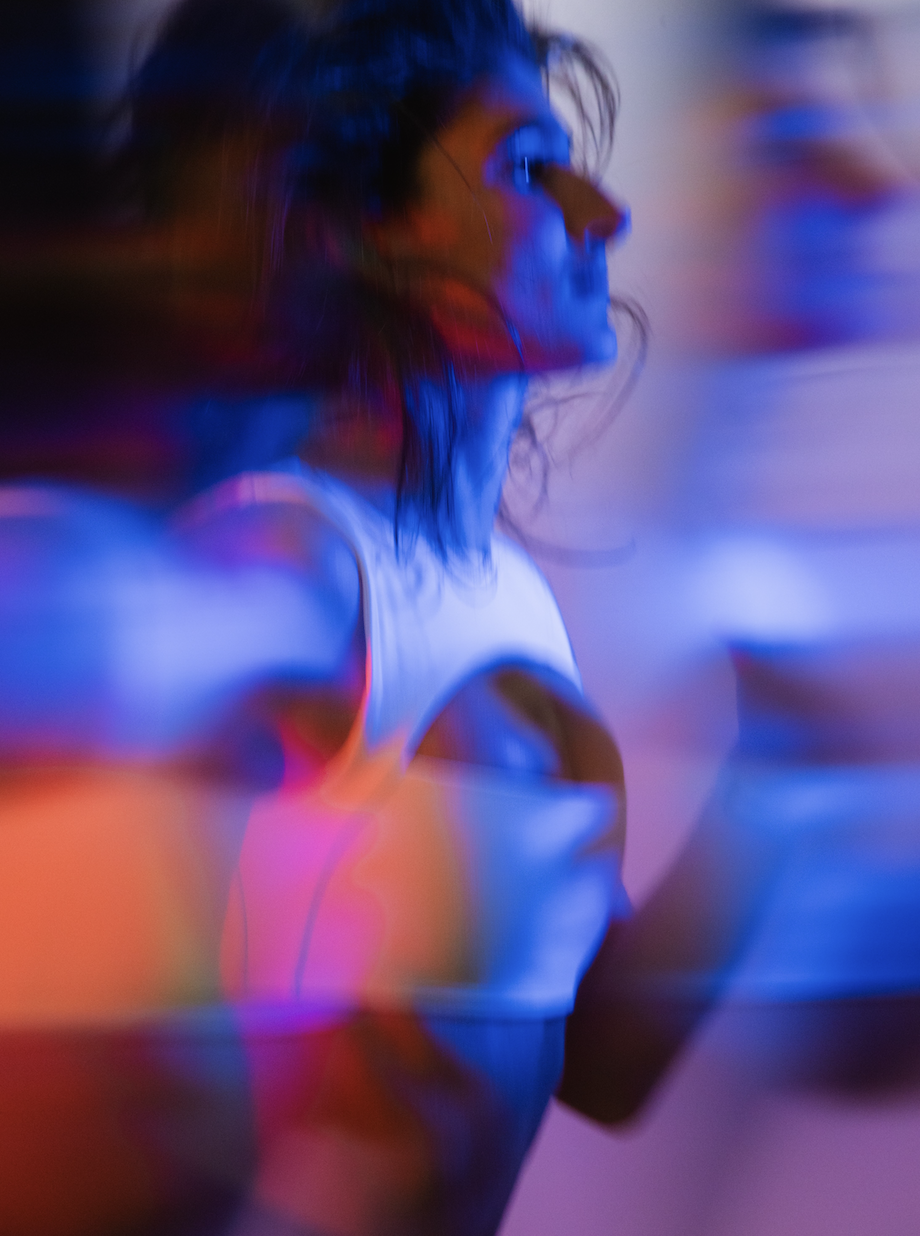 A woman with glasses and long hair, wearing a white tank top, in a blurry, colorful environment with blue, purple, and red lighting.