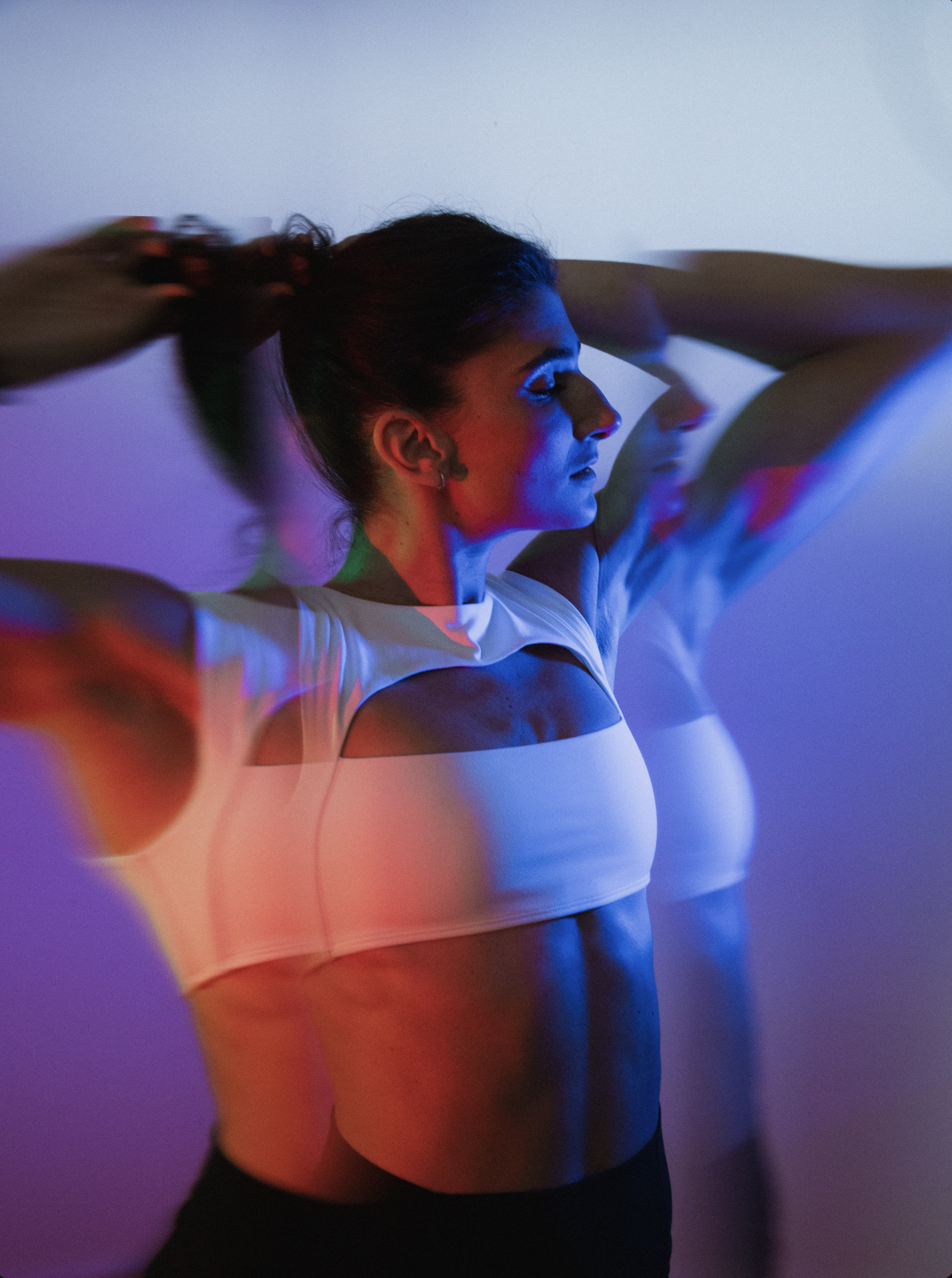 A woman with dark hair tied in a ponytail, wearing a white crop top, is seen posing against a wall with multicolored lighting, creating a distorted impression of her reflection.