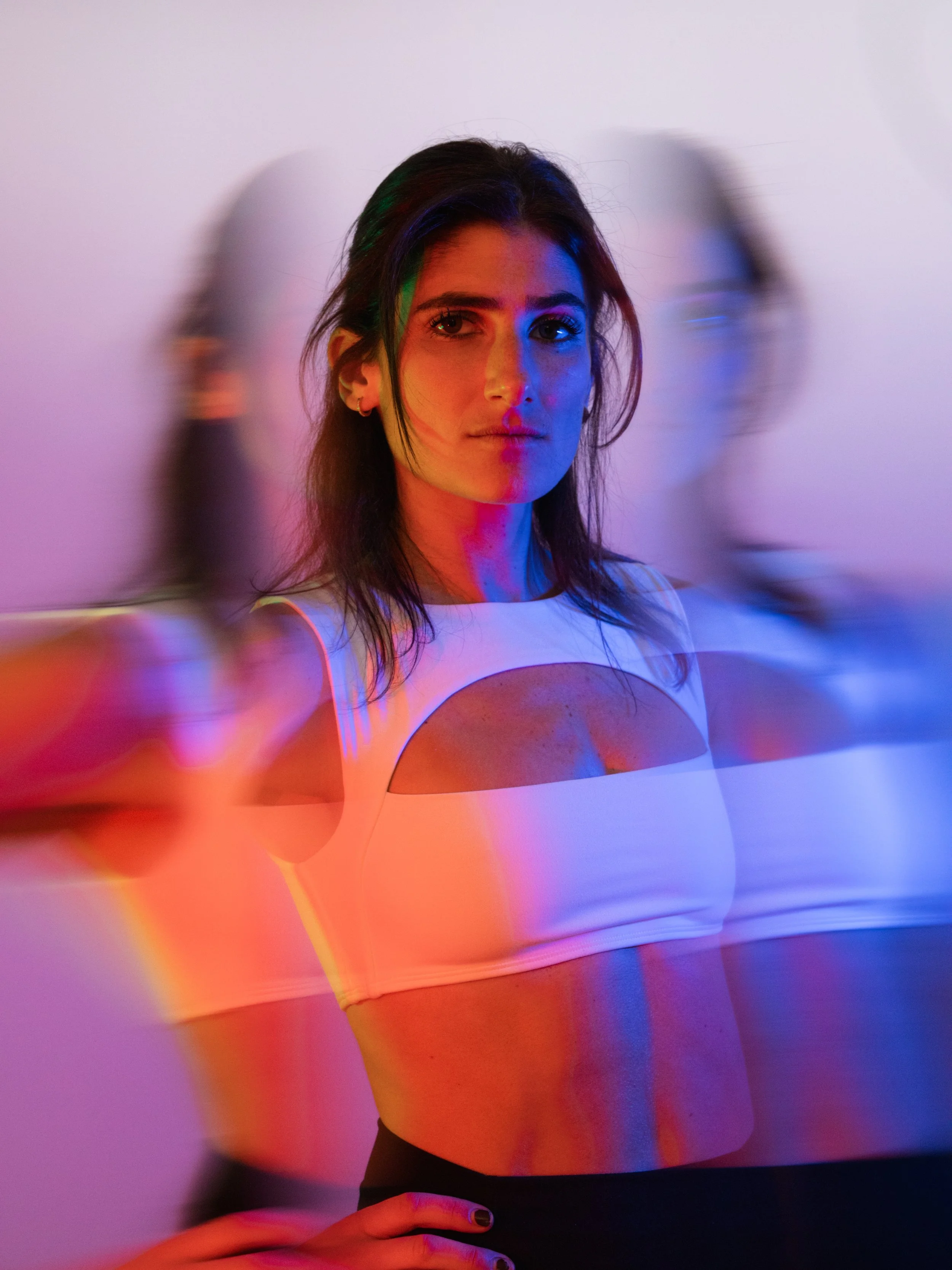 A woman with dark hair and light skin is standing in front of a white background. The photo has colorful lighting effects creating a rainbow-like reflection on her face and clothing. Two blurred figures are visible on either side of her.
