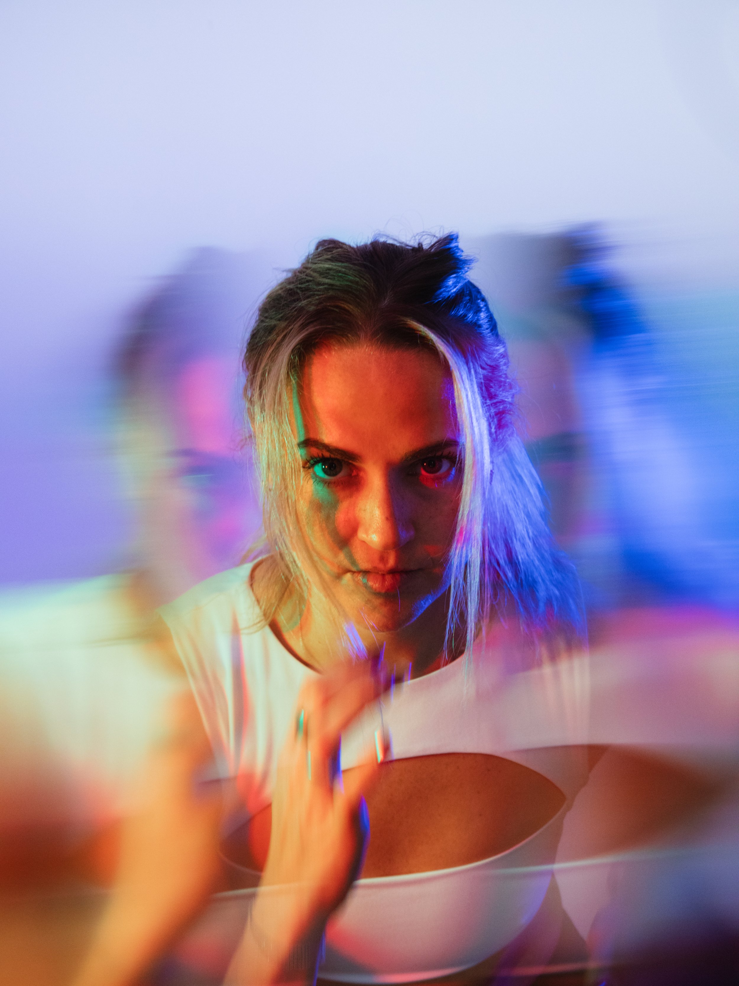 A woman with blonde hair styled in a messy bun and wearing a white top. She is making direct eye contact with the camera with a serious expression. The background features colorful, blurred light effects creating a psychedelic and vibrant atmosphere.