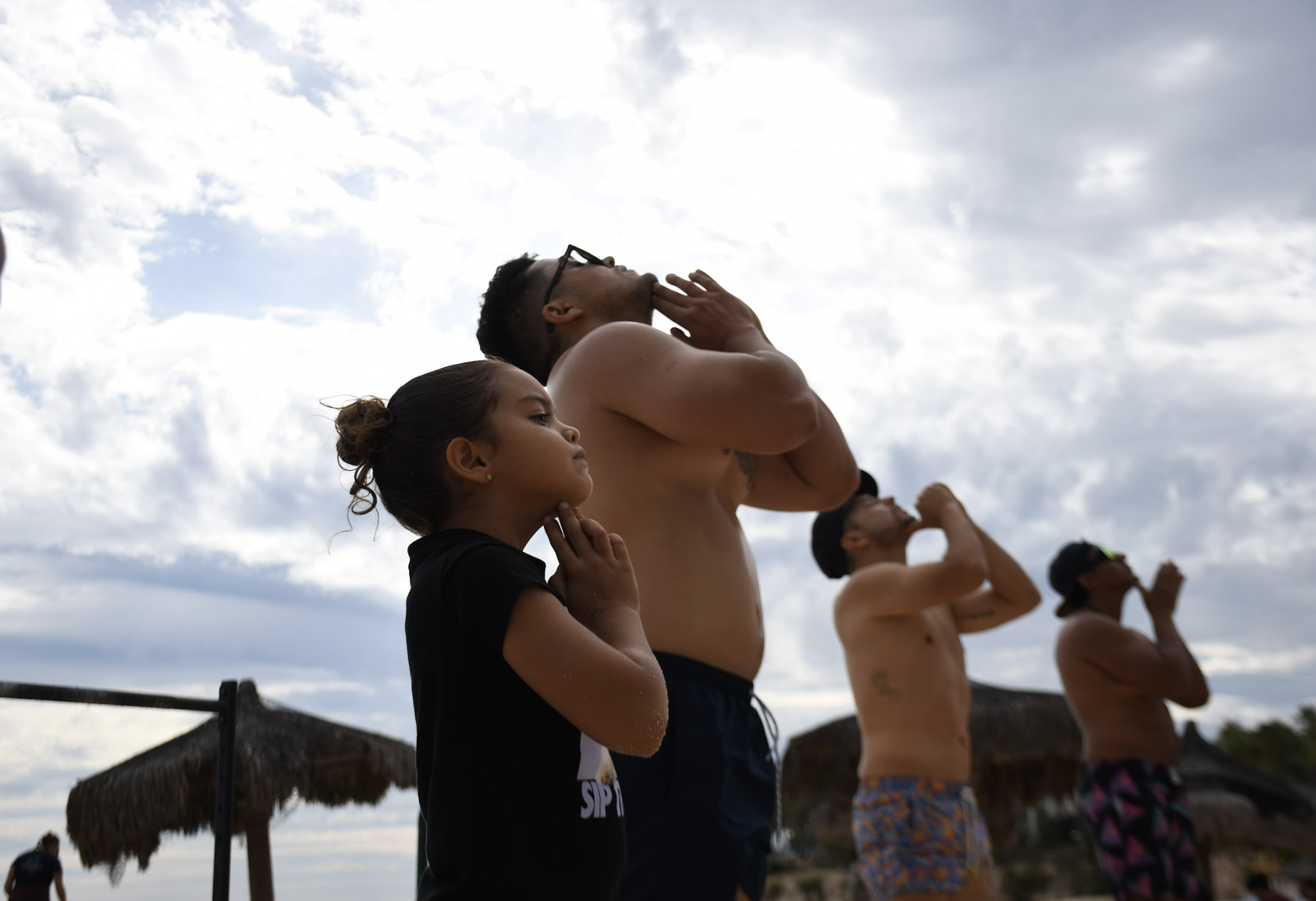 Cuatro personas en la playa, con los ojos cerrados y manos en la garganta, en posición de meditación o yoga, con nubes en el cielo y sombrillas de paja en el fondo.