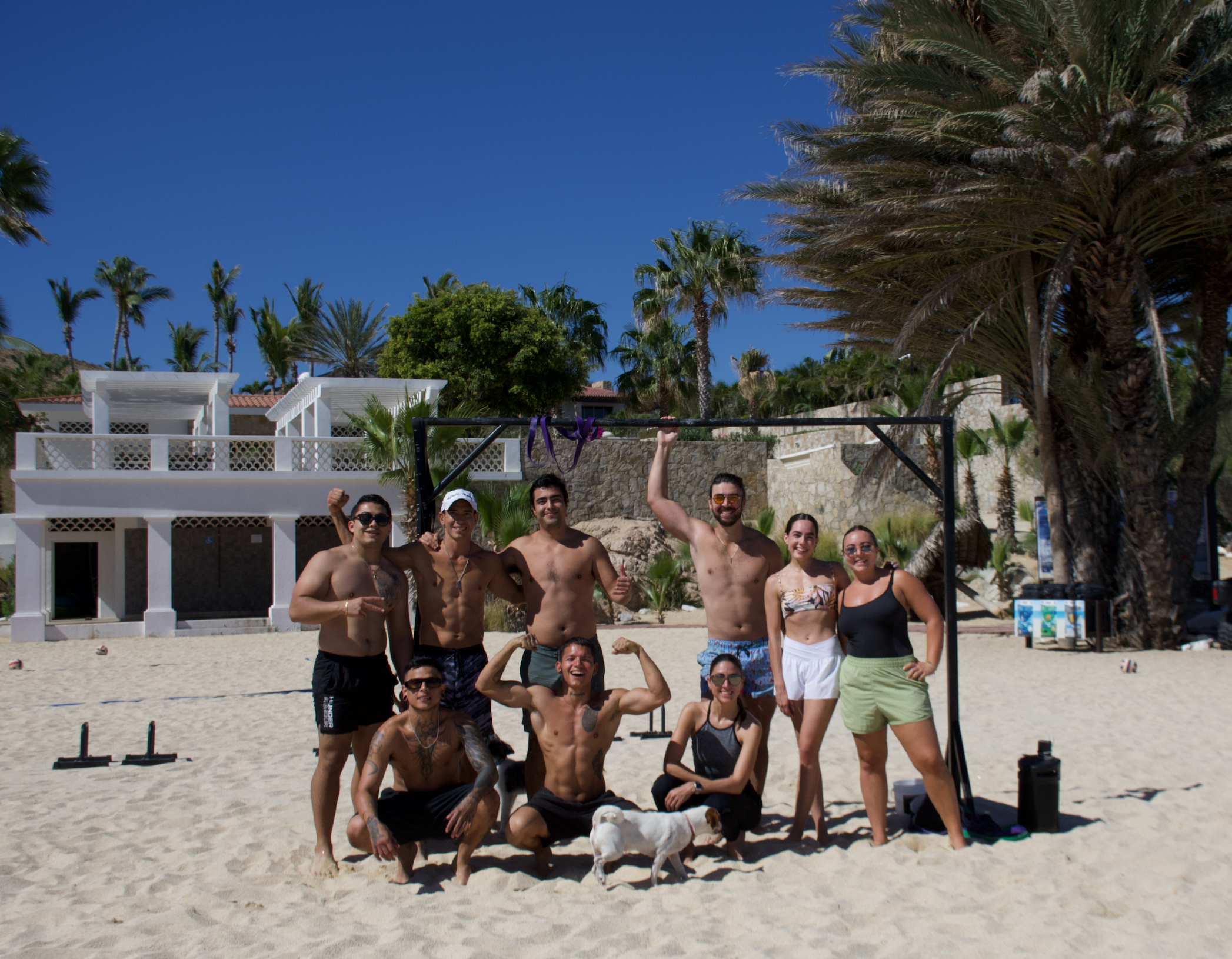 Grupo de personas en la playa posando frente a un arco de entrenamiento, algunos levantando los brazos y sonriendo, con palmeras y edificios blancos de fondo, en un día soleado.