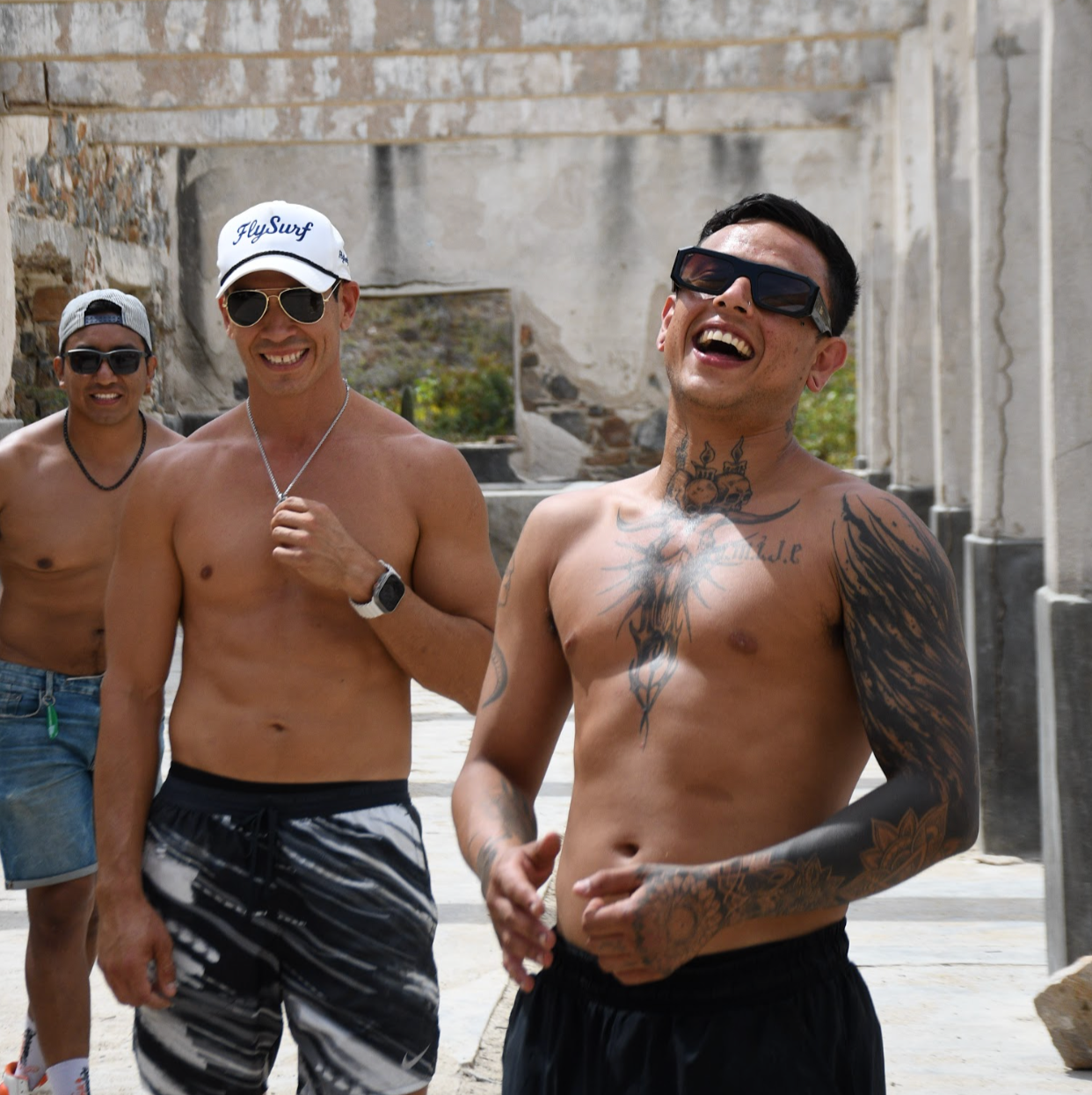 Cuatro hombres sin camiseta y usando gafas de sol riendo y disfrutando al aire libre, con una estructura en ruinas de fondo.