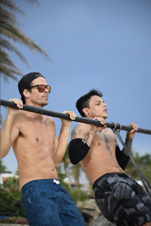 Dos hombres sin camiseta usando gafas y pantalones cortos, uno con tatuajes y piercing en la oreja, sosteniendo una barra en un entorno de playa.