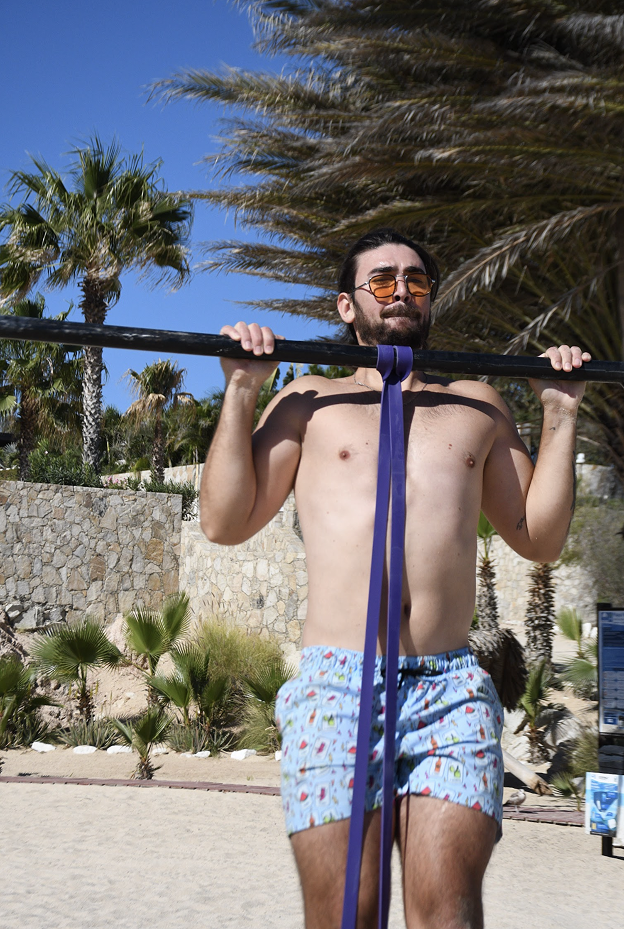 Hombre sin shirt con gafas de sol sosteniendo una barra con una banda azul en un entorno de playa con palmeras y cielo despejado.
