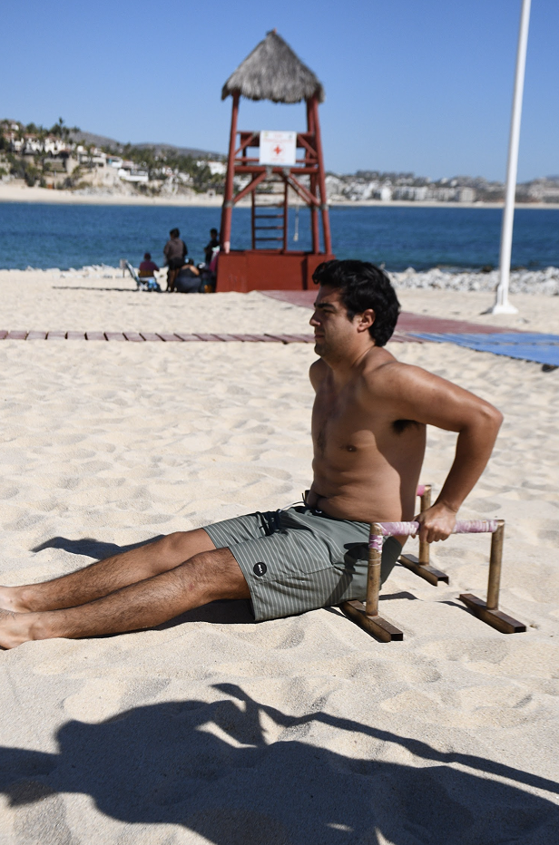 Hombre con camiseta de baño haciendo ejercicio en la playa, sentado en un aparato de entrenamiento de arena, con mar, sombrilla de palapa, y estructura de salvavidas en el fondo.