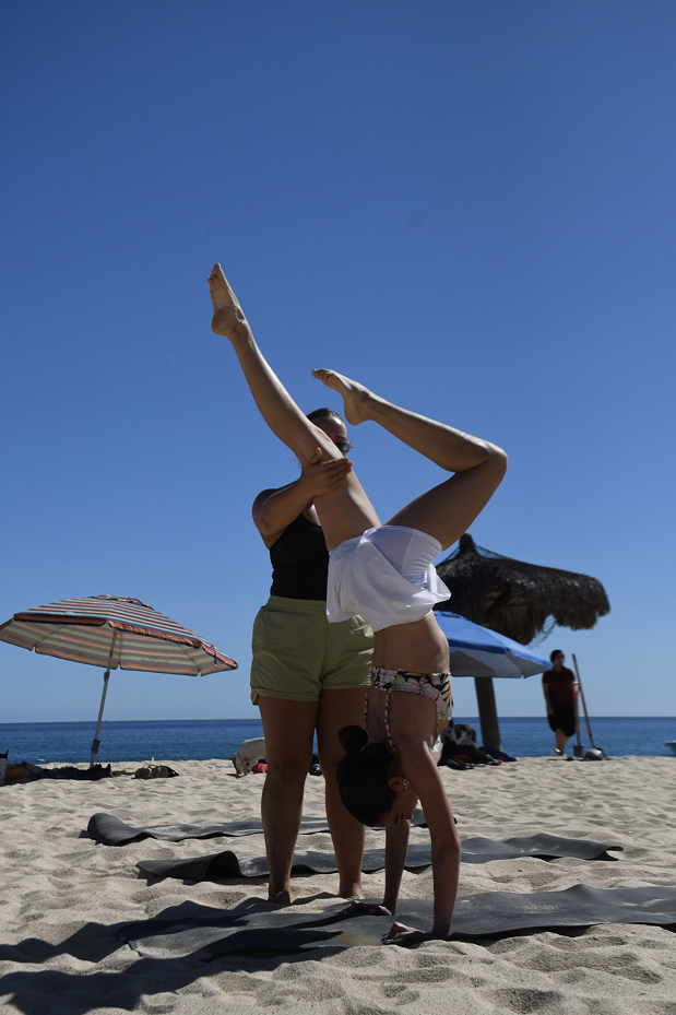 Dos mujeres en la playa, una ayudando a la otra a hacer una acrobacia de yoga en la arena, con sombrillas y palapas en el fondo y el mar bajo un cielo despejado.