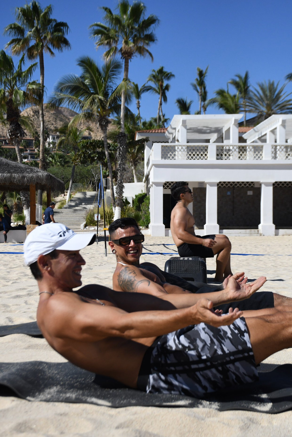 Personas descansando en la playa, algunos riendo y disfrutando del sol, con palmeras y construcciones blancas de fondo.