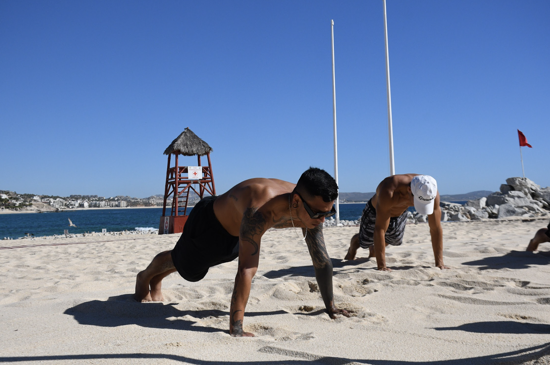 Dos hombres haciendo ejercicio en la playa bajo el sol, en posición de plancha, con fondo de mar y estructuras de bandera y torre de vigilancia.