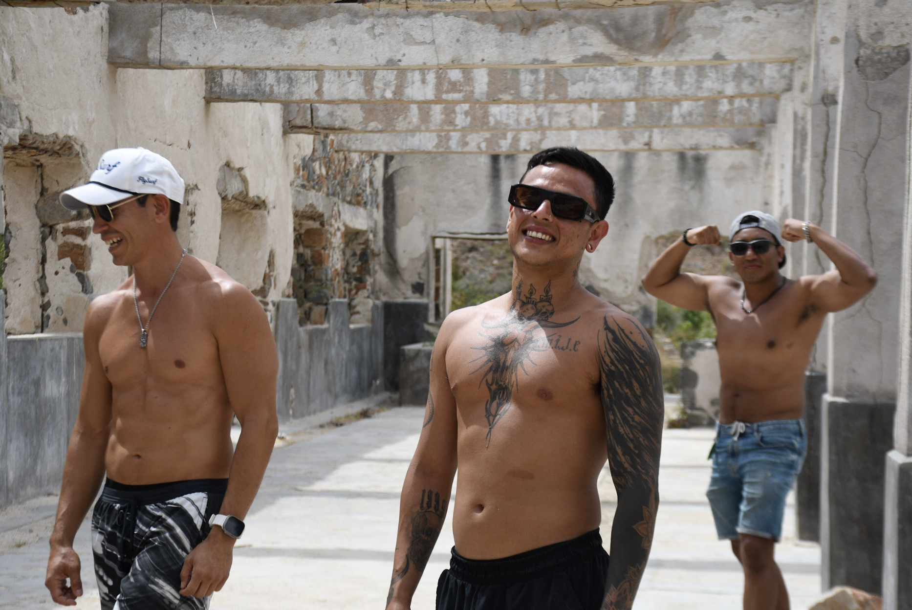 Tres hombres semidesnudos con gafas de sol en un lugar al aire libre con paredes ruinosas y estructuras en ruinas, sonriendo y mostrando músculos.