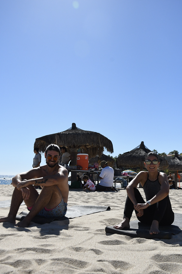 Dos personas sentadas en la arena de la playa con sombrillas de paja, disfrutando del día soleado.