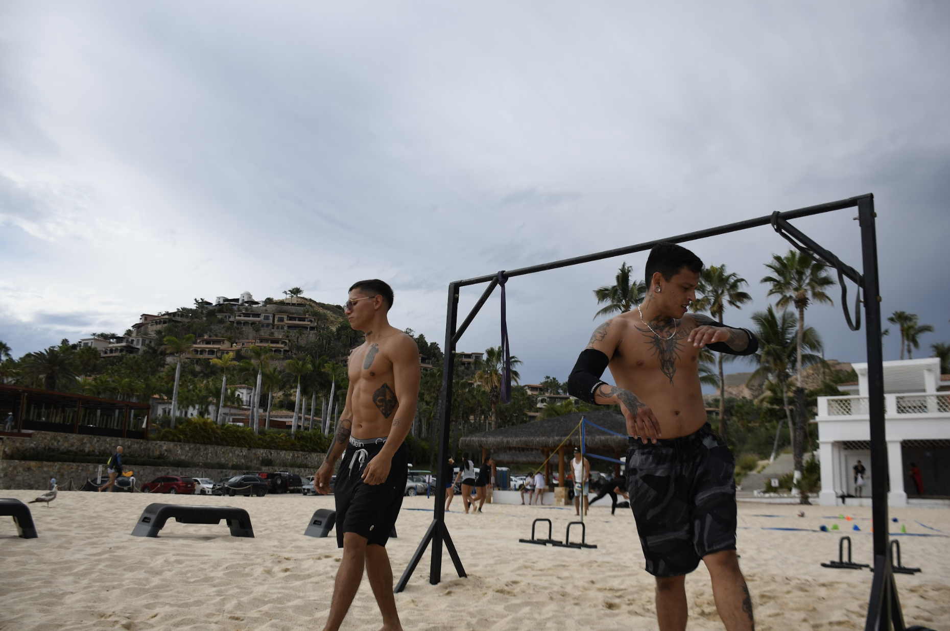 Dos hombres sin camisa en la playa, uno de ellos ajustándose la ropa y el otro mirando hacia el lado, con palmeras y casas en las colinas y un cielo nublado en el fondo.