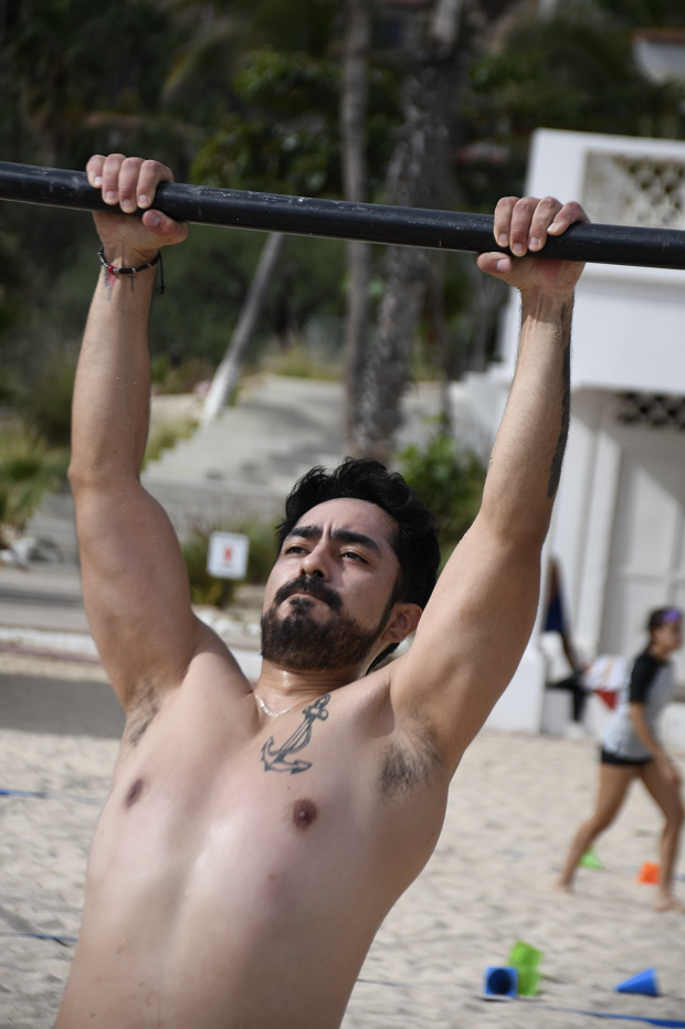 Hombre calvo y con barba realizando ejercicio de tirón en una playa, sin camiseta, con tatuaje de ancla en el pecho, fondos de árboles y estructuras de playa.
