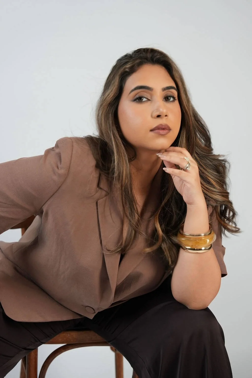 A woman with long wavy brown hair wearing a brown blazer and black pants, sitting on a wooden chair against a plain white background, with her hand resting under her chin, looking at the camera.
