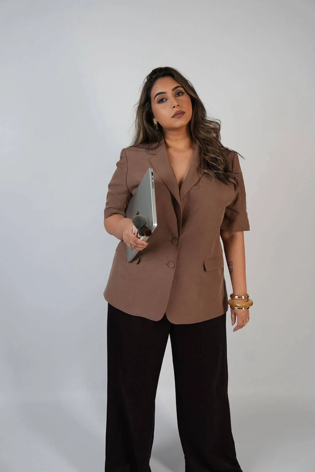 Professional woman with dark wavy hair, wearing a brown blazer and black pants, holding a laptop and a microphone, standing against a plain white wall.