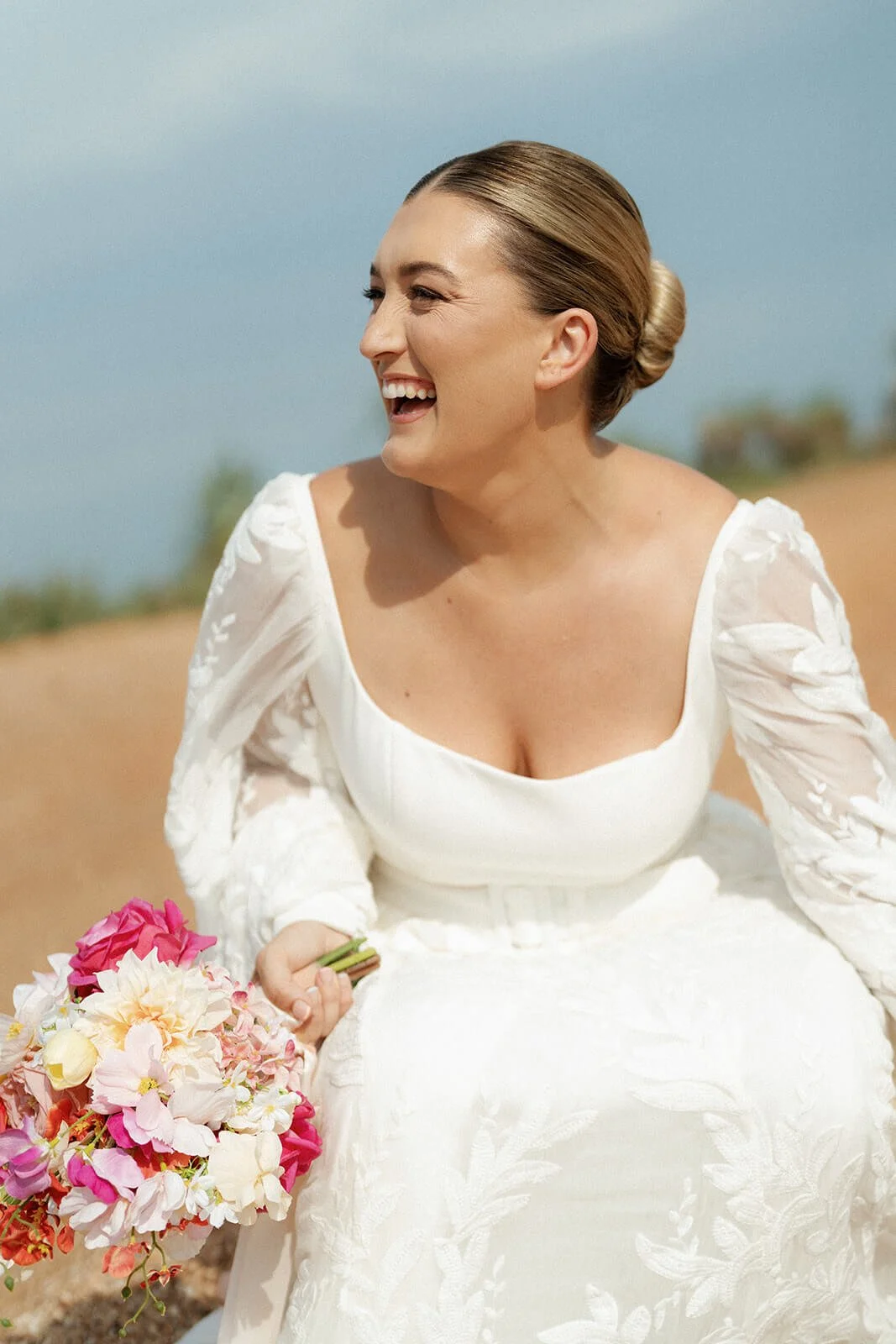 A woman in a white dress holding a bouquet of pink and white flowers, smiling and squinting against the sunlight outdoors.
