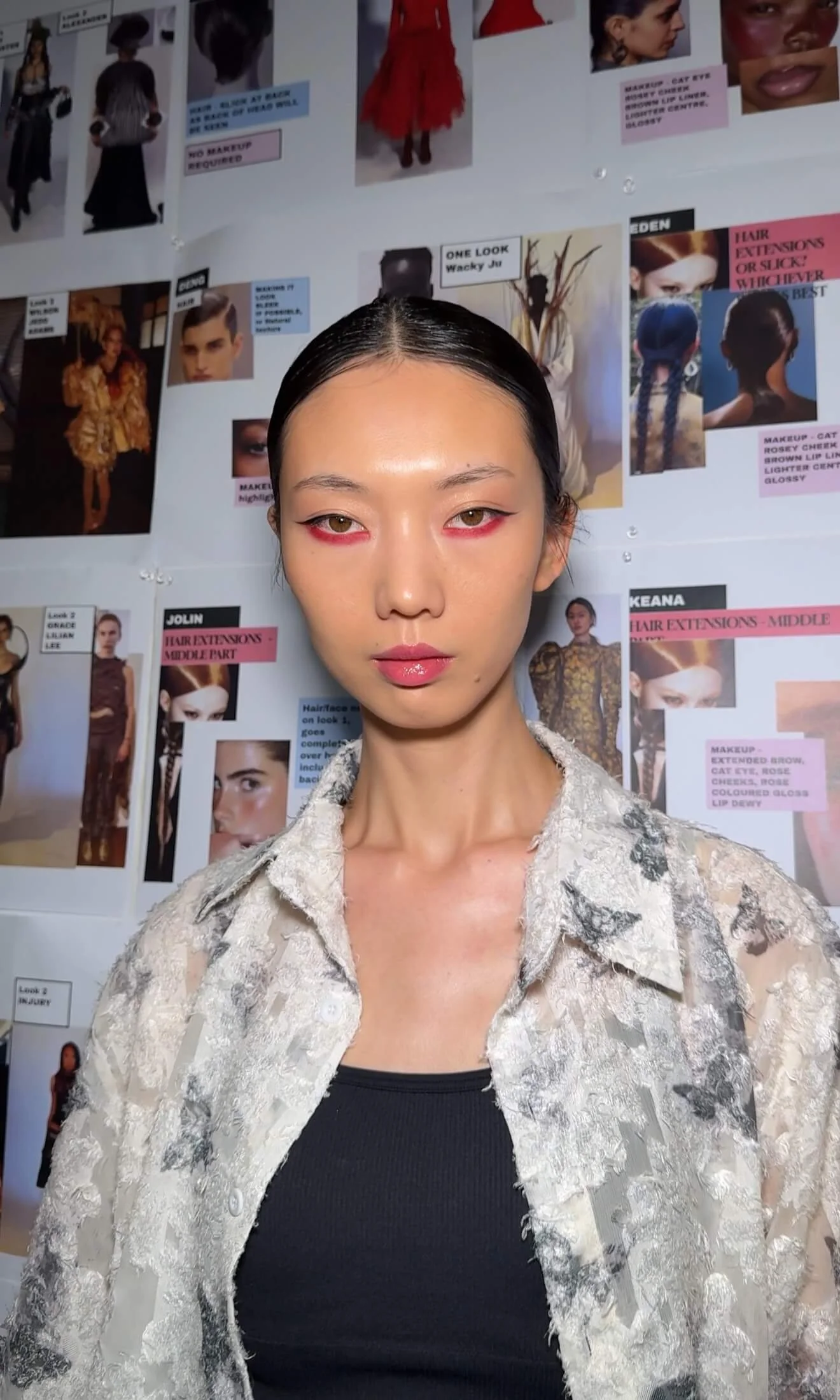 Close-up of a woman with sleek black hair, natural makeup, and pink lip gloss, standing in front of a white wall decorated with fashion magazine pages and photos.