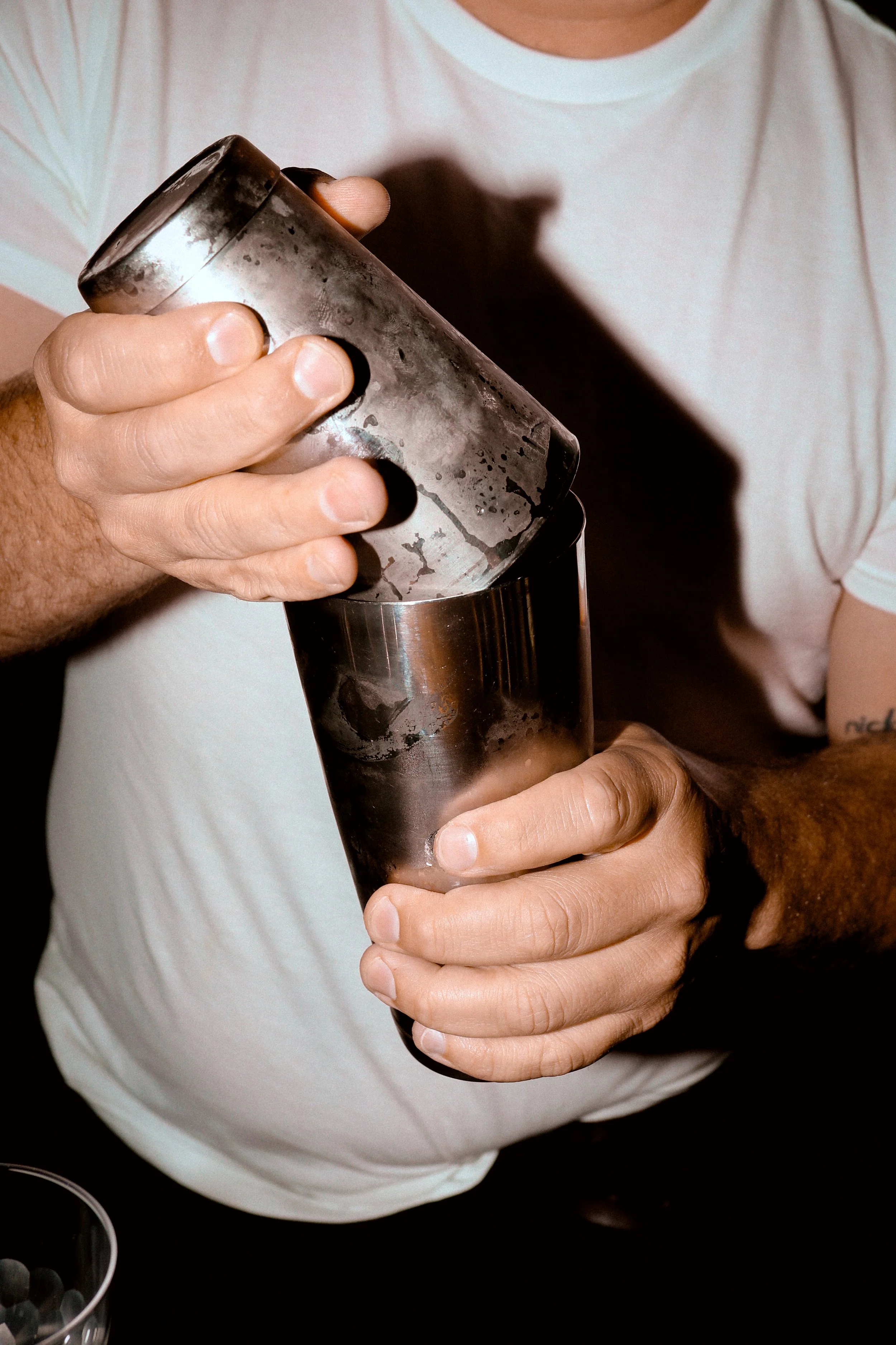 Close-up of a person's hand holding a cocktail shaker with ice, pouring a drink into a glass. The person is wearing a white shirt.