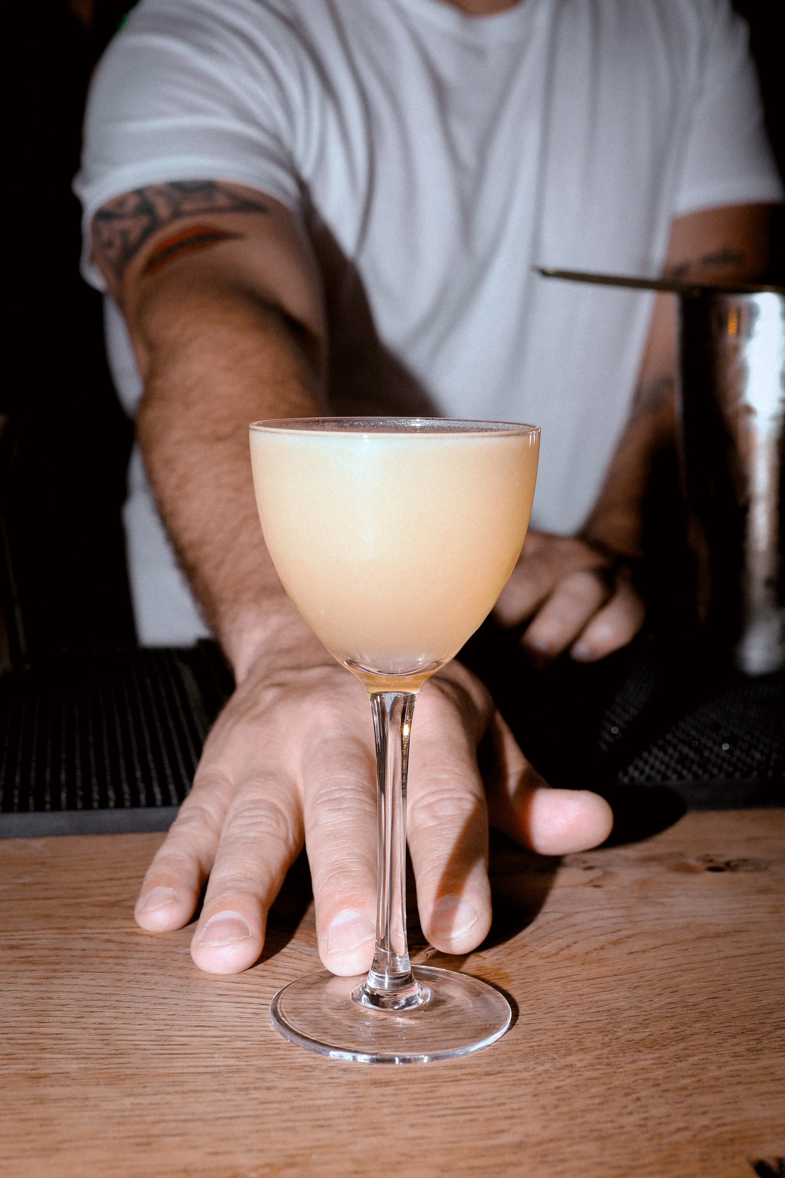 A person in a white shirt with tattoos on their arms is reaching forward with their hand on a wooden table. In front of the person, there is a tall, frosted glass with a light-colored drink.
