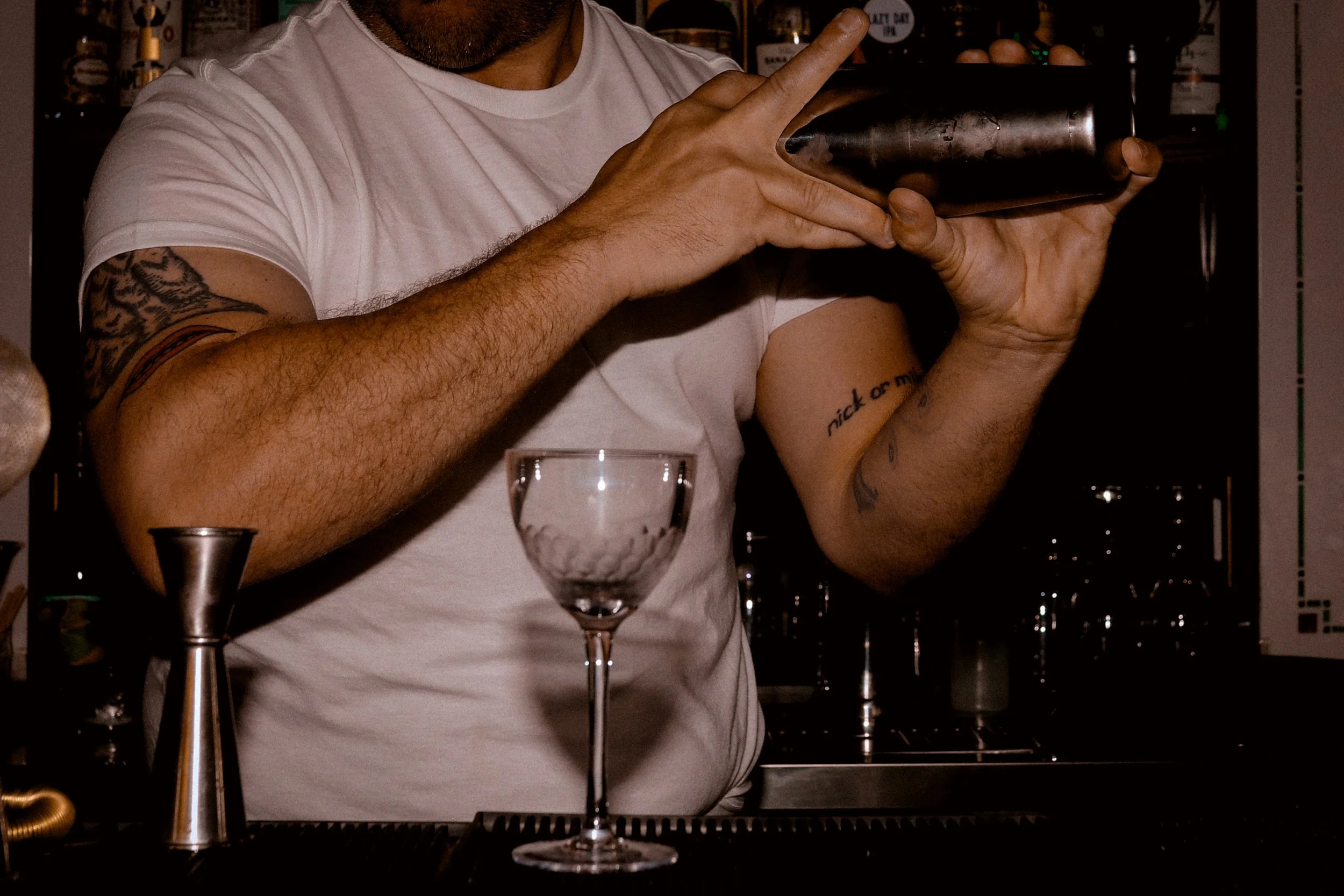 A person with tattoos on his left arm and a tattoo on his right wrist, wearing a white t-shirt, is holding a dark metallic cocktail shaker in a bar environment. Several bottles are visible on shelves behind him. In front of him, there is a long-stemm