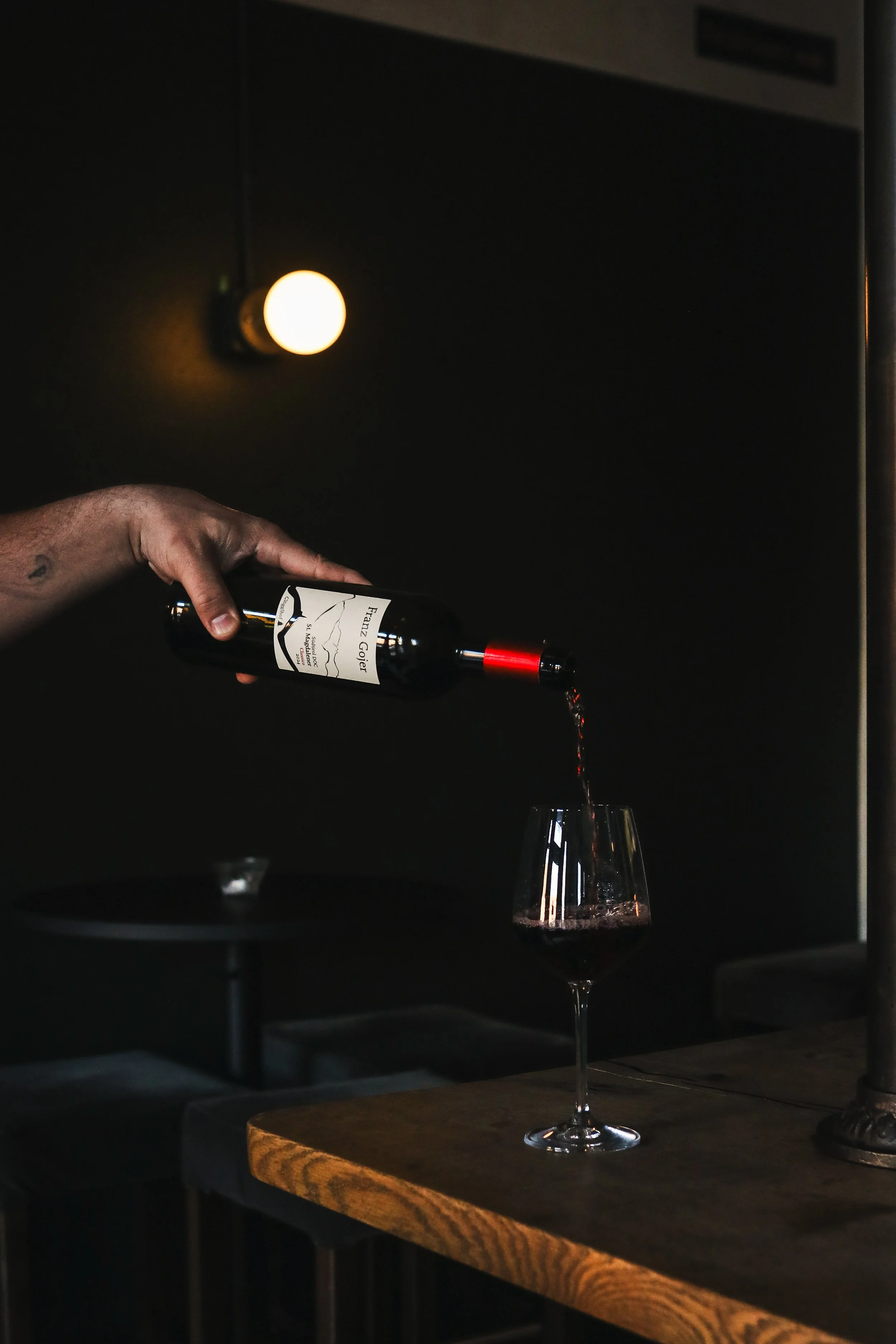 A hand holding a wine bottle pouring red wine into a glass on a wooden table, with a dark background and a warm light source.
