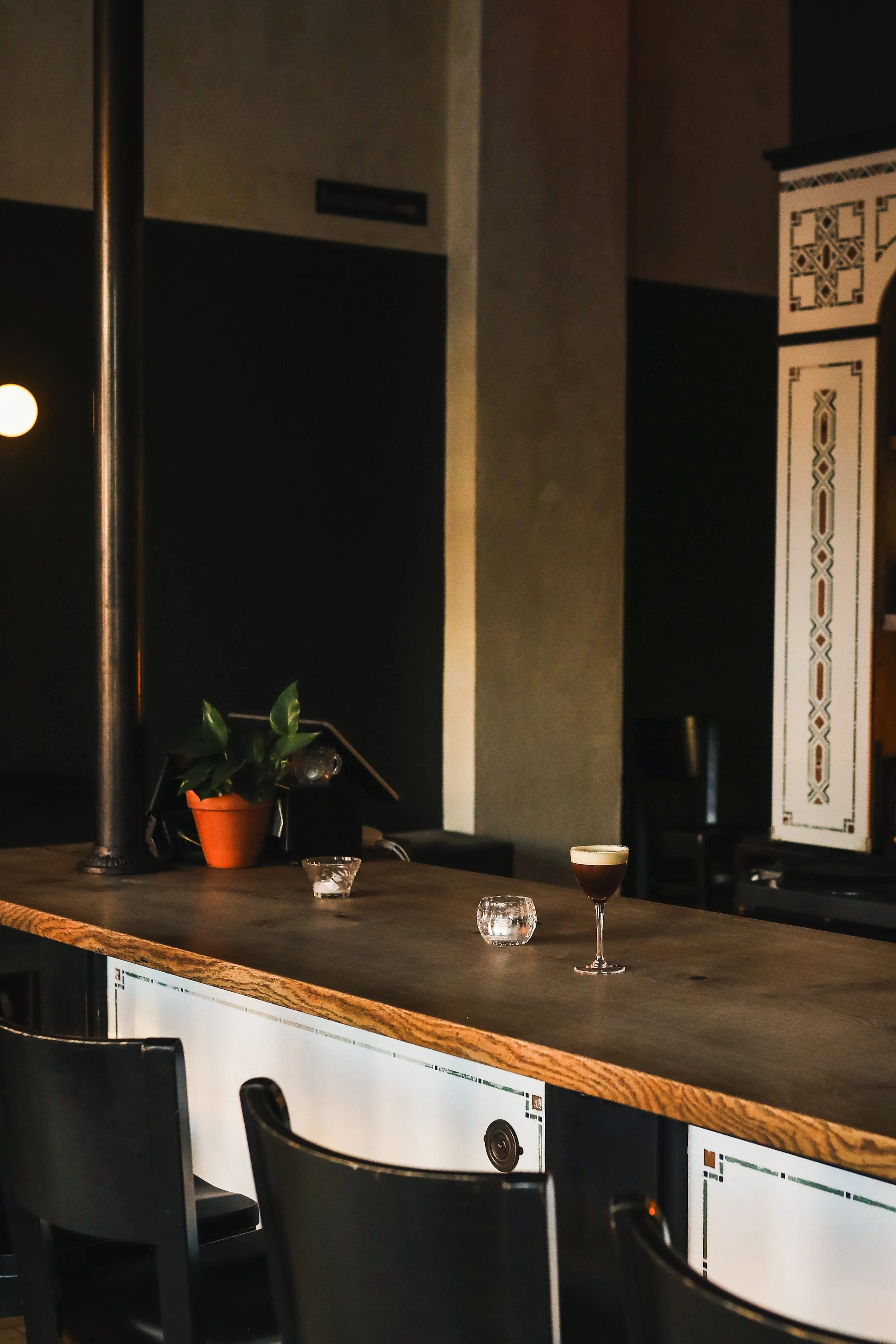 Interior of a bar or café with a wooden countertop, glassware, a potted plant, and a drink with foam, with dark walls and decorative paneling.
