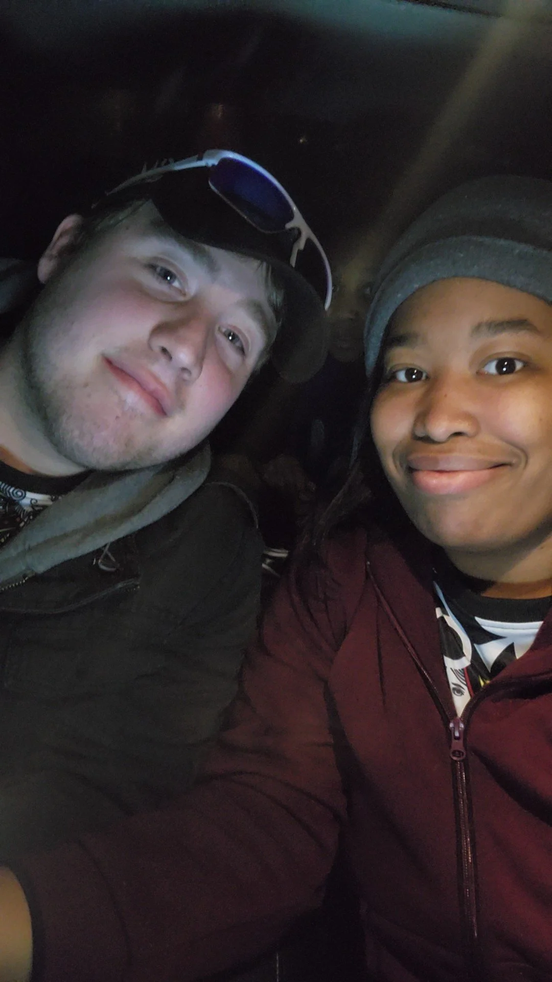 Two young couple smiling for a selfie in a dimly lit setting, one wearing a black jacket and sunglasses on his head, the other wearing a maroon jacket and a grey beanie.