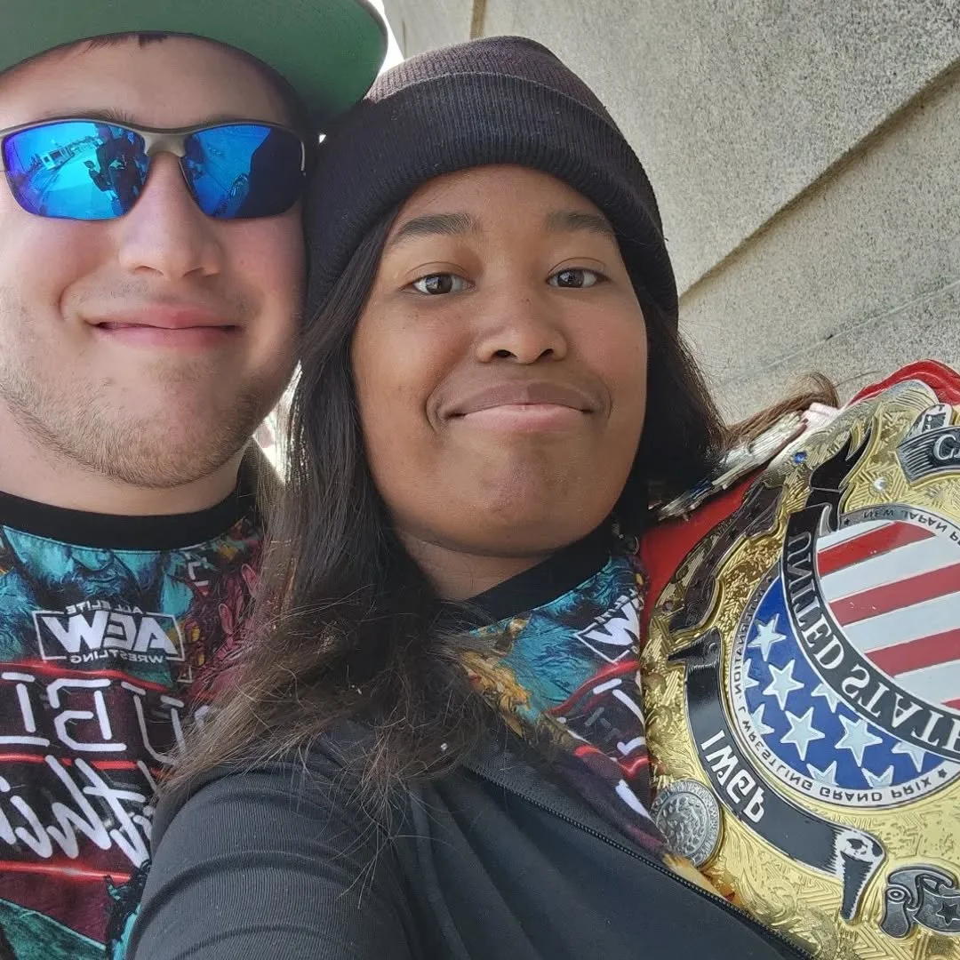 Two smiling men taking a close-up selfie, wearing colorful shirts and accessories, with one holding an championship belt.