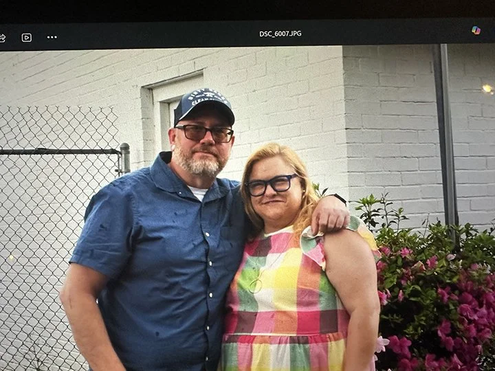 A man and a woman standing next to each other outdoors, with the man wearing a cap and glasses, and the woman wearing glasses and a colorful dress, in front of a white brick wall and flowering bushes.