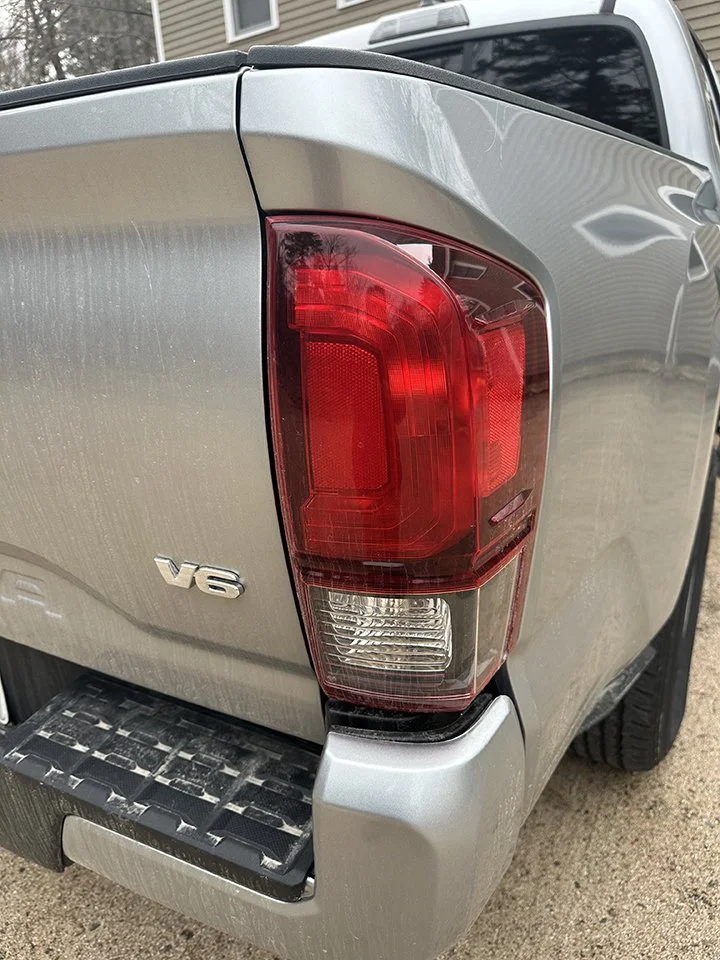 Close-up of the rear left corner of a silver pickup truck, showing the tail light, V6 badge, and tailgate with step bumper.